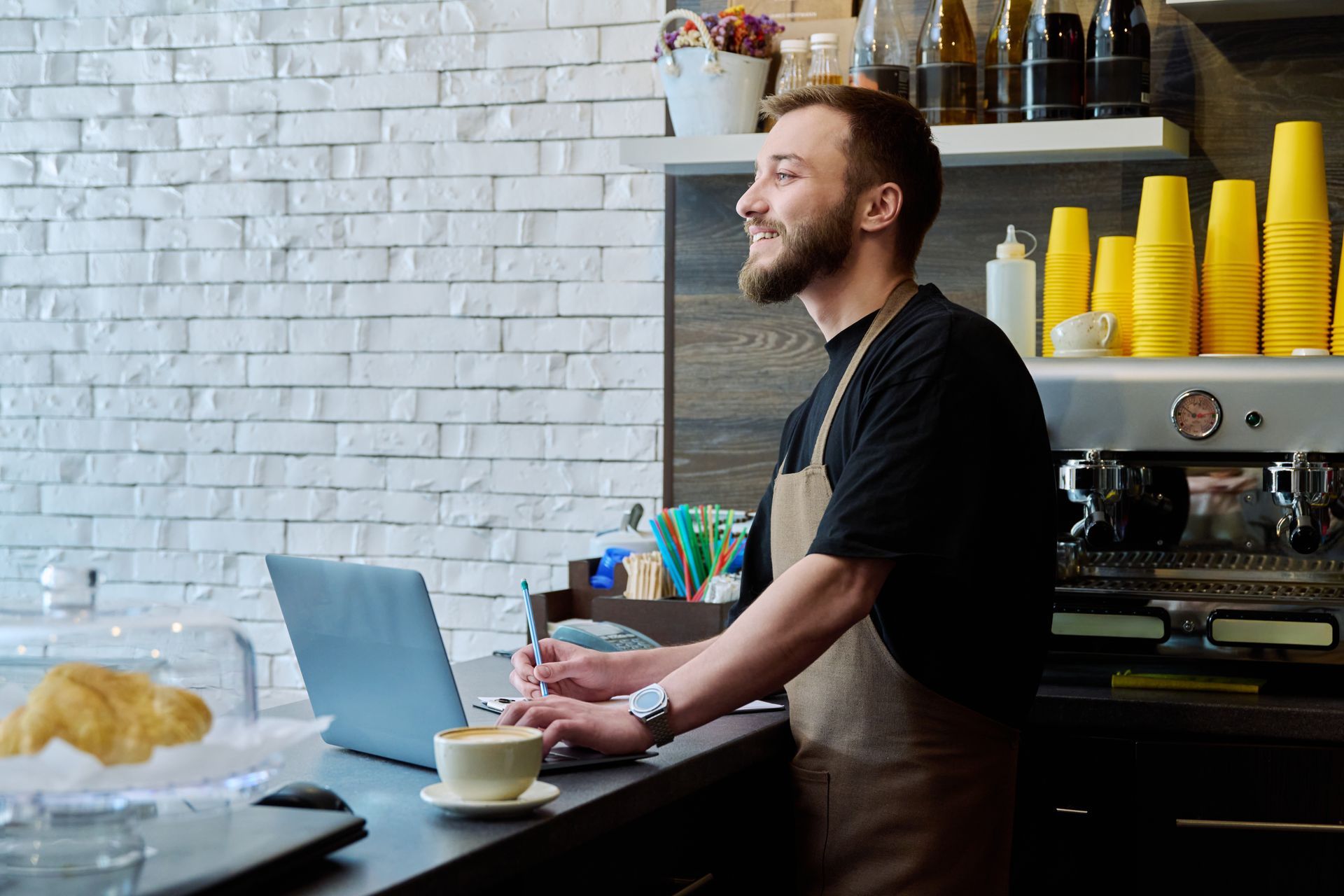 Barista using laptop and writing, smiling, at cafe counter. Brick wall background.
