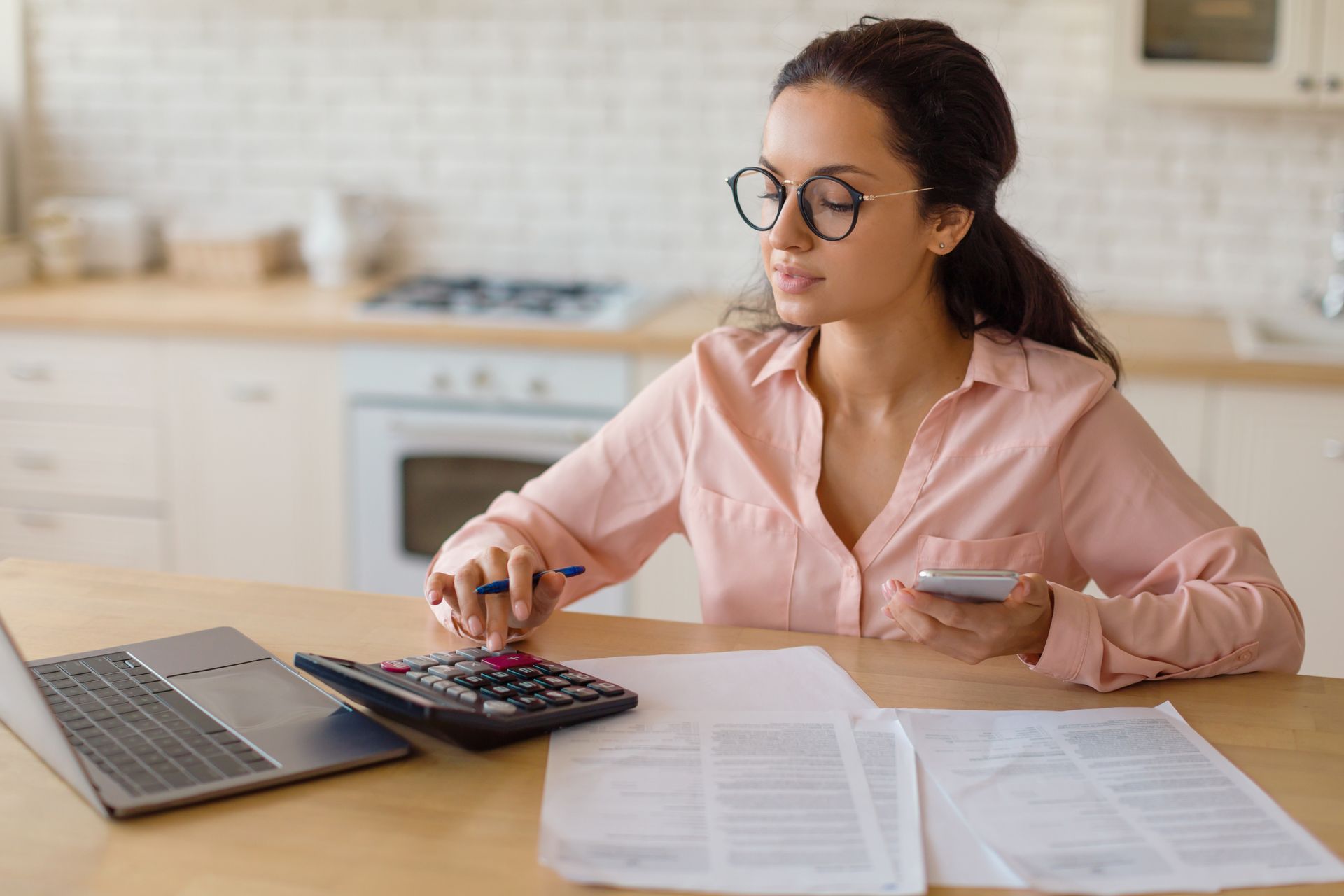 Woman with glasses using a calculator, phone, and laptop in a kitchen, reviewing documents.