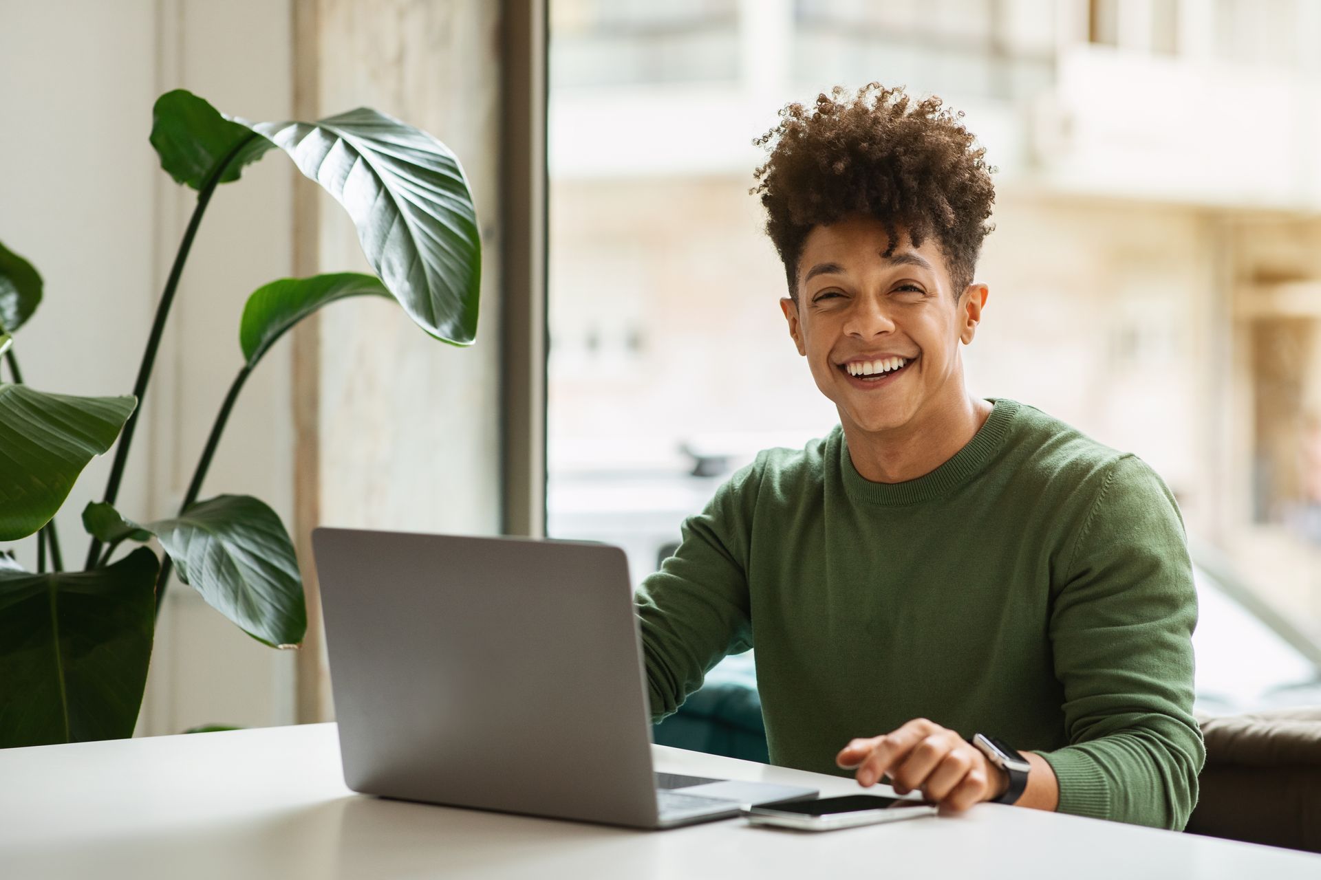 Person smiling at laptop indoors, green sweater, plant, window.