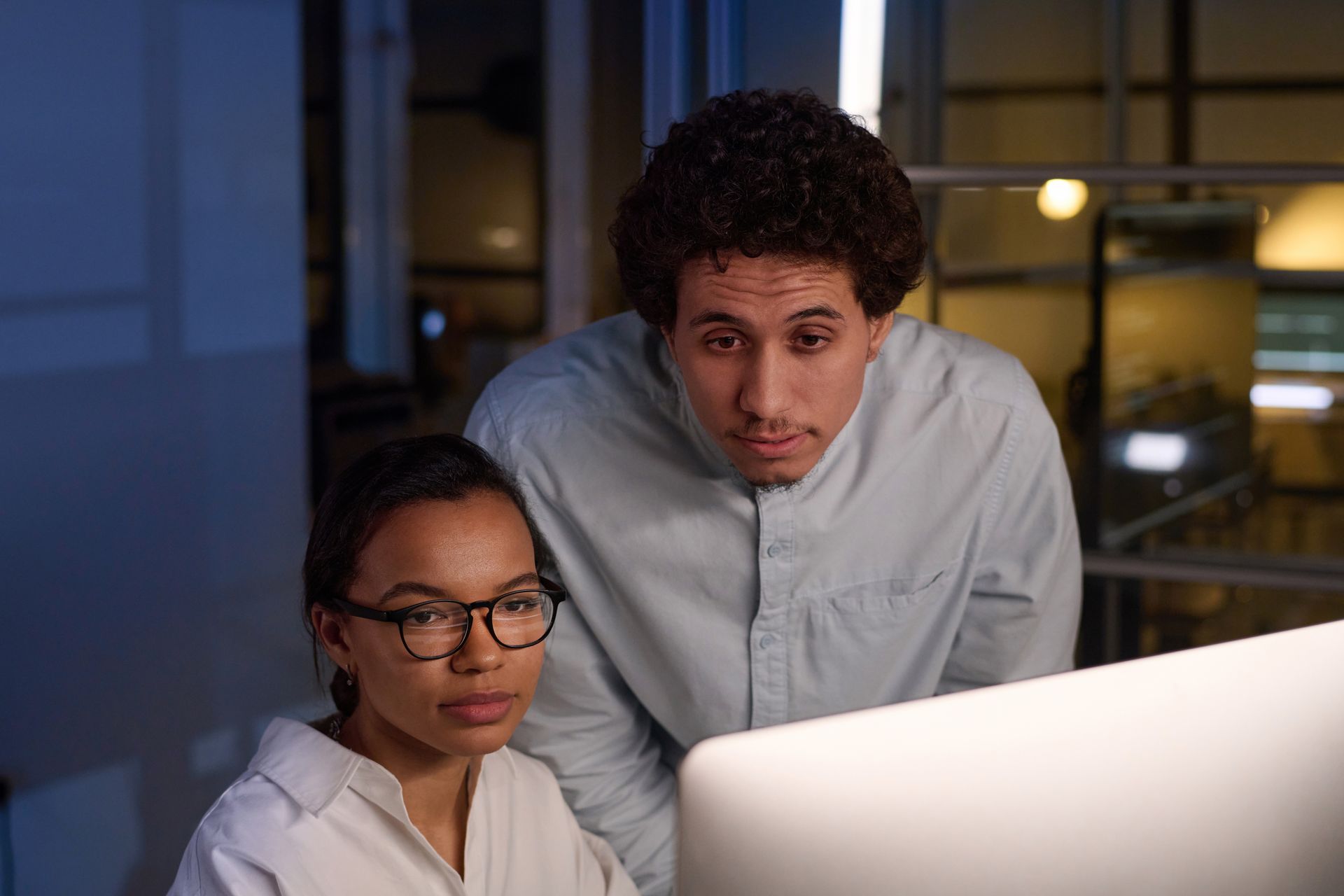 Woman and man looking at a computer screen together, focused. Low-light office setting.