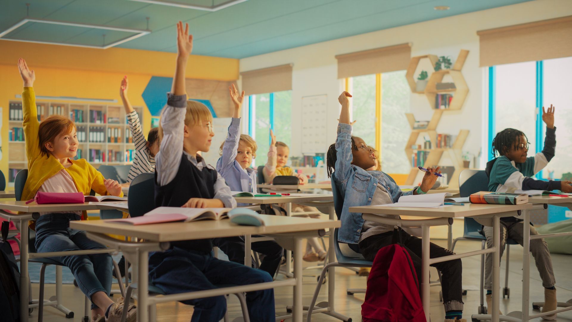 Children raising hands in a classroom, answering a question. Sunny, bright classroom.