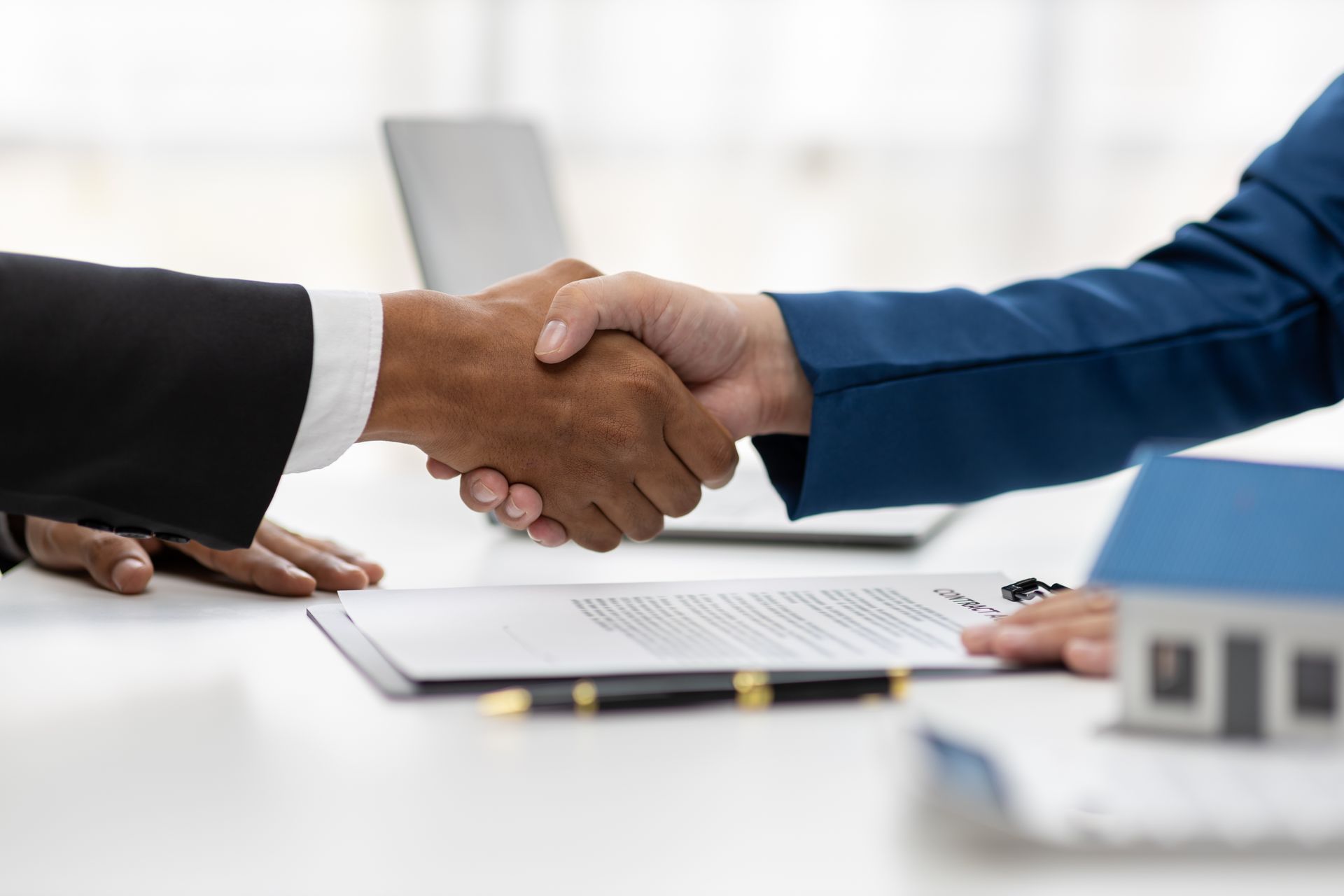 Two people shaking hands over a contract, a small house, and laptop on a desk.