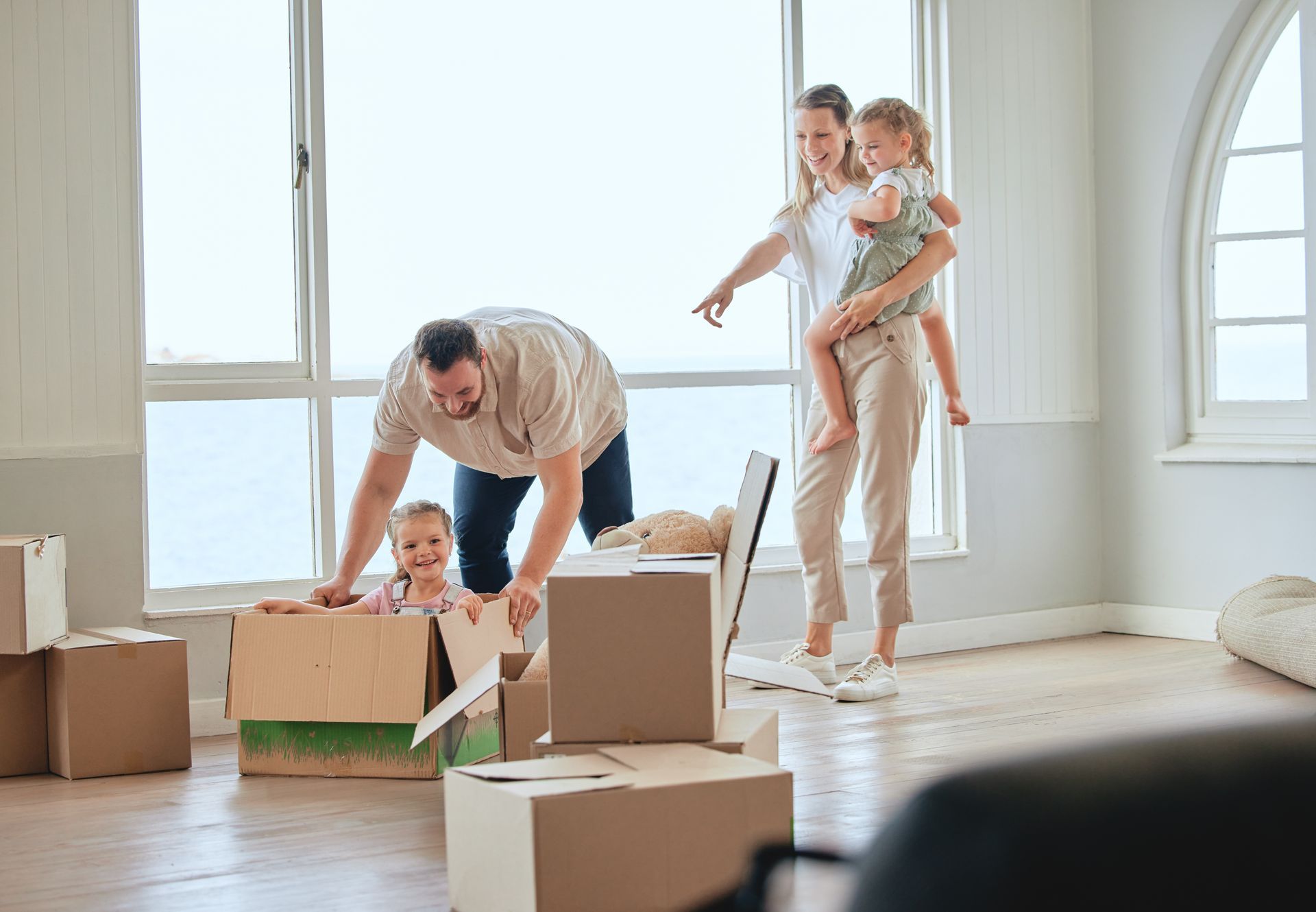 Family unpacking in new home with cardboard boxes. Man helping child in box, woman holding child near windows.