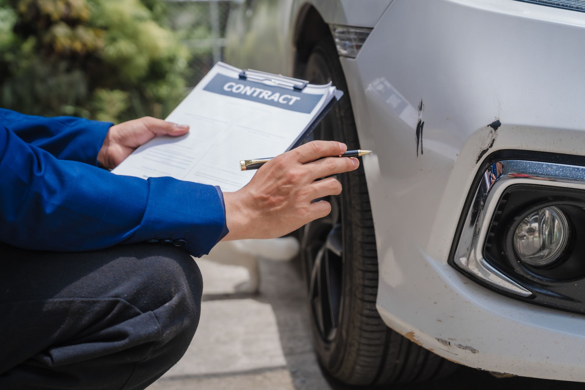 Person inspecting damage on a white car, holding a contract on a clipboard.