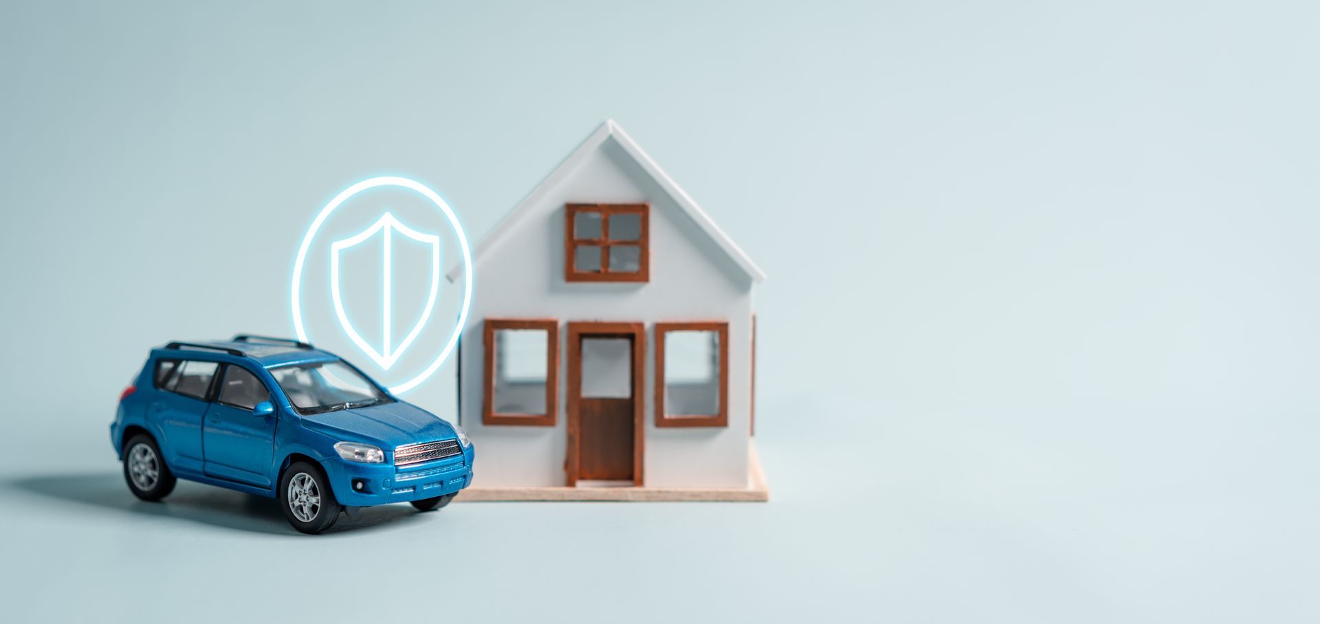 Blue car and toy house with shield icon, symbolizing insurance, on light blue background.
