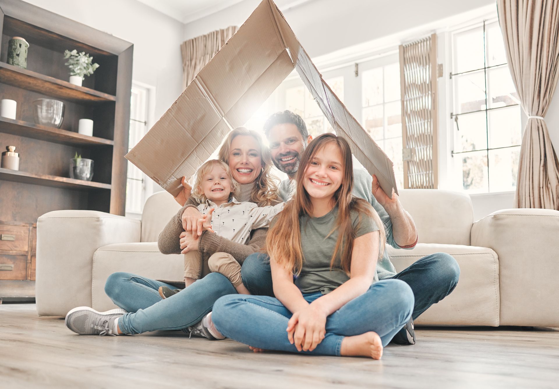 Family under a cardboard house roof, smiling in a living room with sunlight.