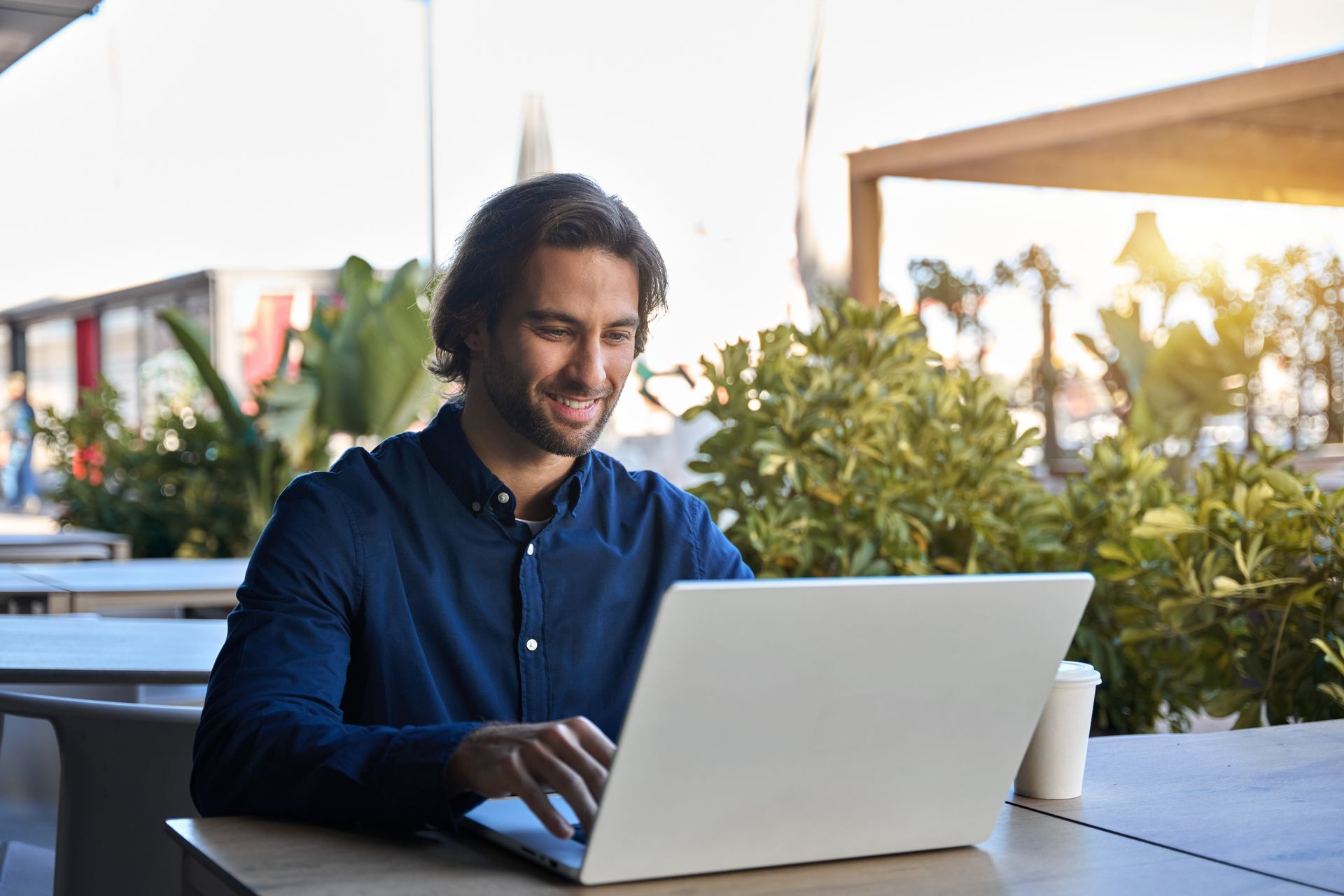 Man using a laptop at an outdoor table, smiling. Blue shirt, coffee cup, and greenery in the background.