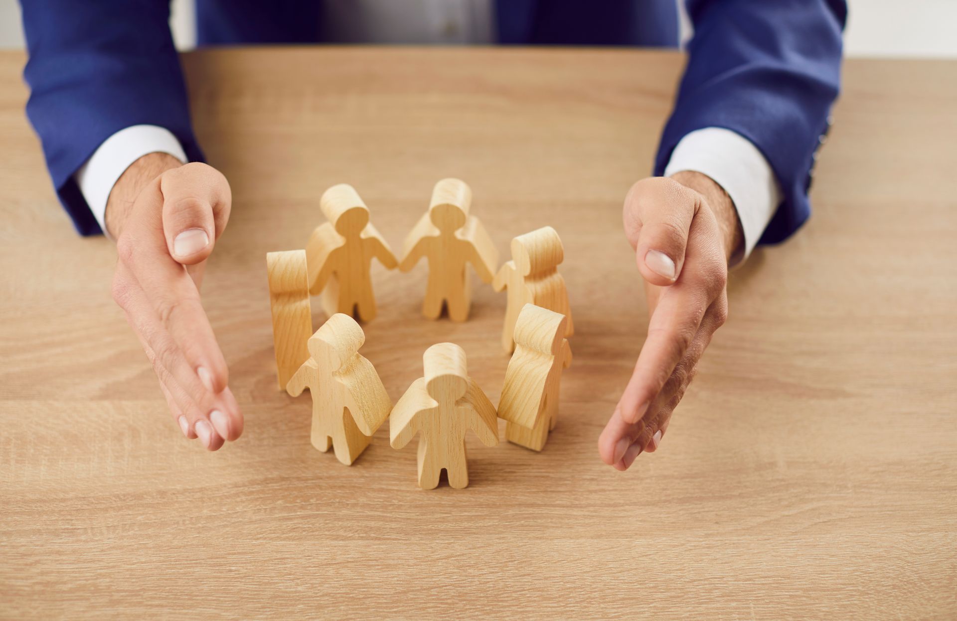 Hands of a person in a suit protecting a circle of wooden figurines on a wooden table.