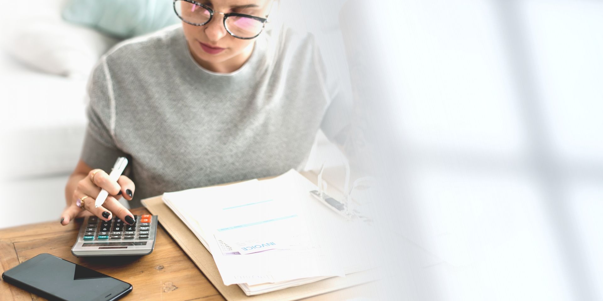 Woman wearing glasses using a calculator and reviewing paperwork on a desk.