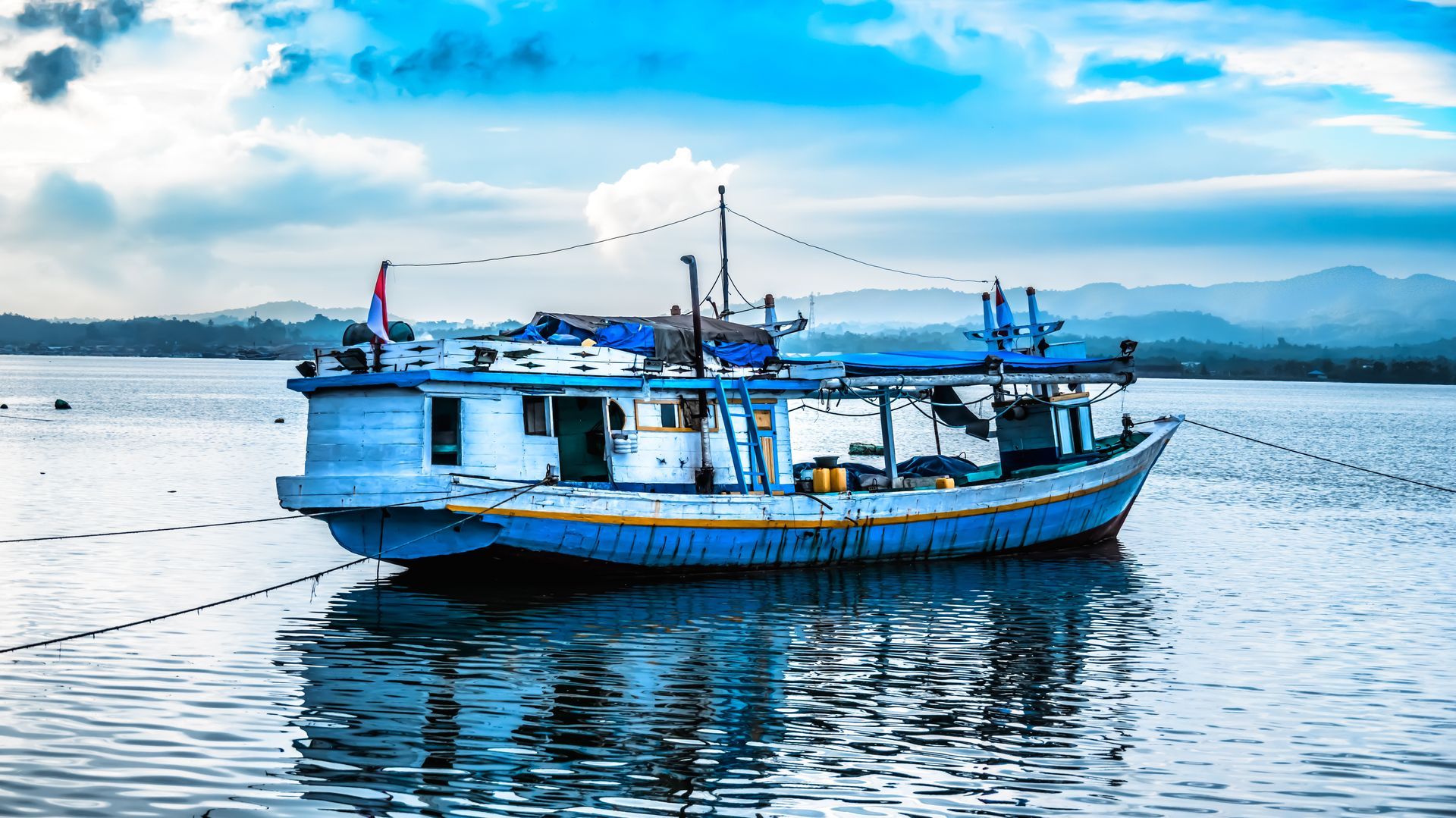 Blue and white fishing boat floating in calm water under a cloudy sky.