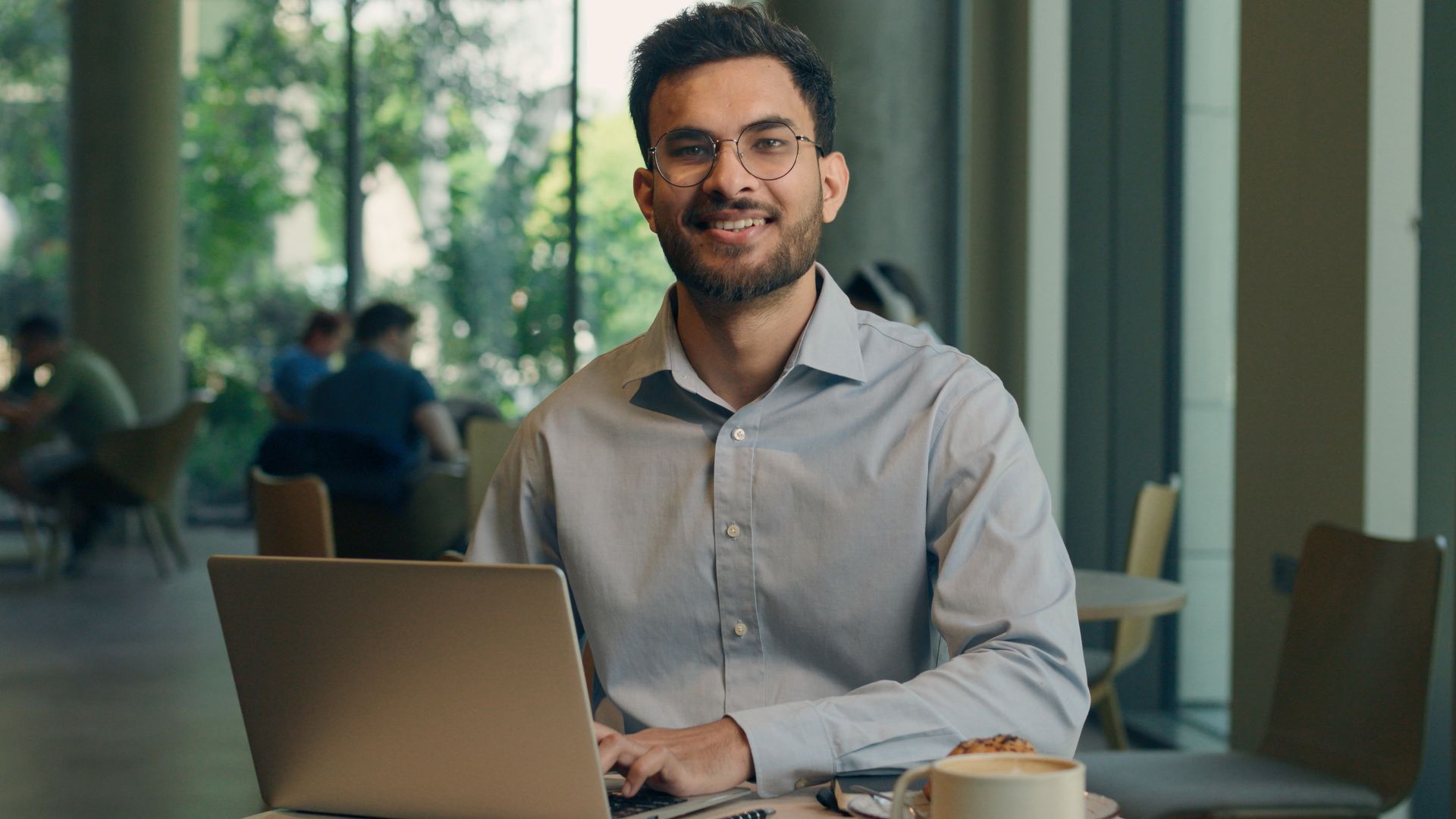 Man wearing glasses smiles at the camera while working on a laptop at a cafe table.