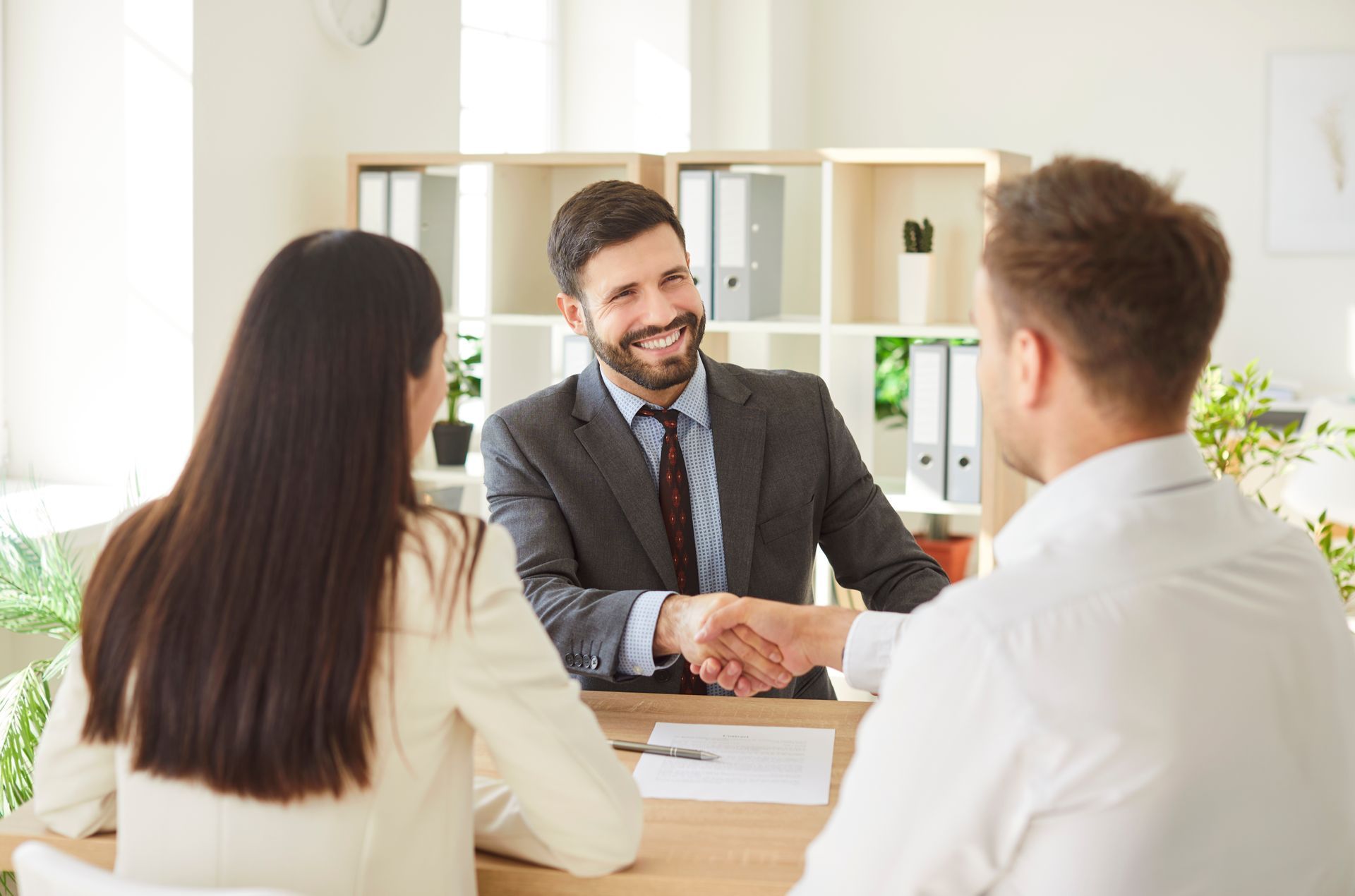 A man shaking hands with another man, seated at a desk, with a woman beside them, in an office setting.