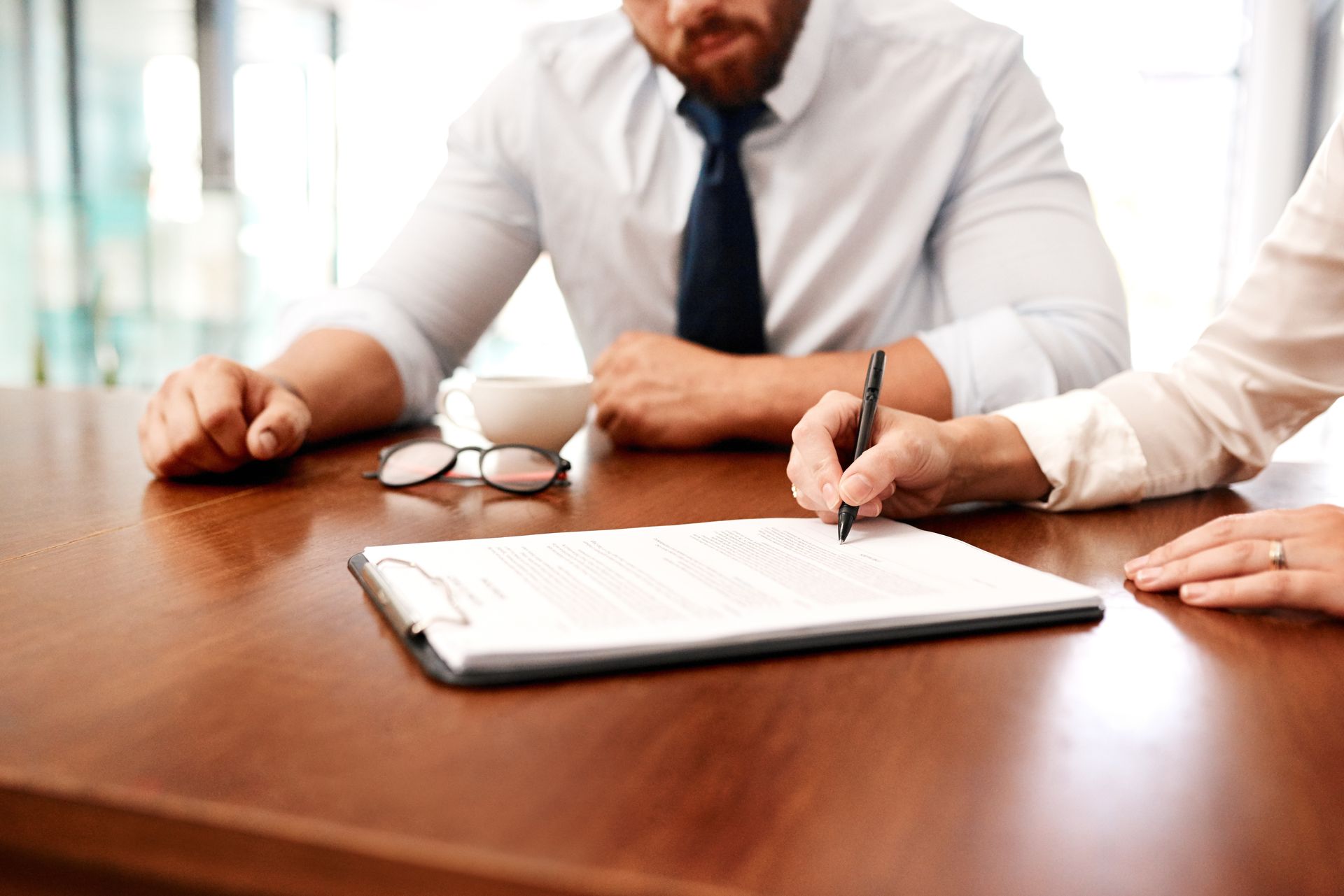 Person signing document on clipboard at wooden table, another person watches. Coffee cup, glasses.