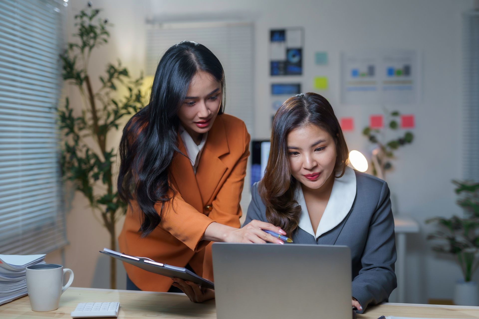 Two women collaborating over a laptop in an office. One points at the screen, holding a clipboard.