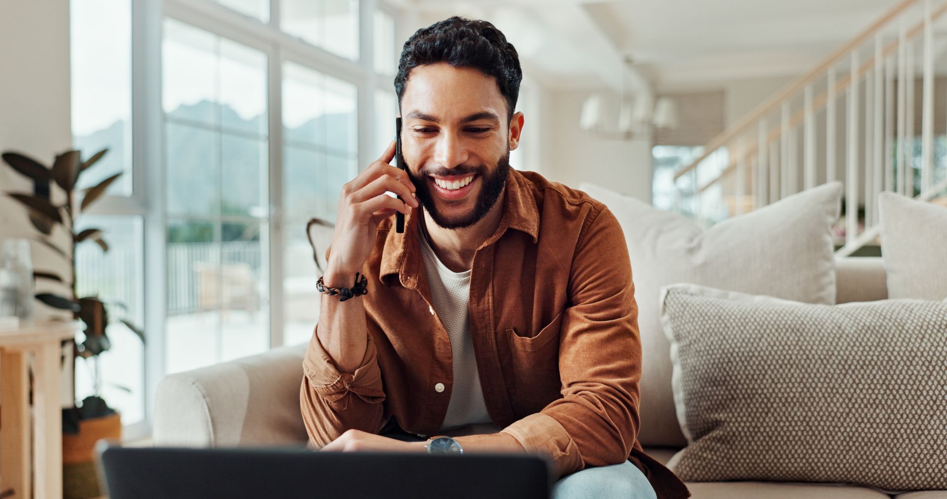 Man smiling while talking on phone, sitting on couch with laptop in bright living room.