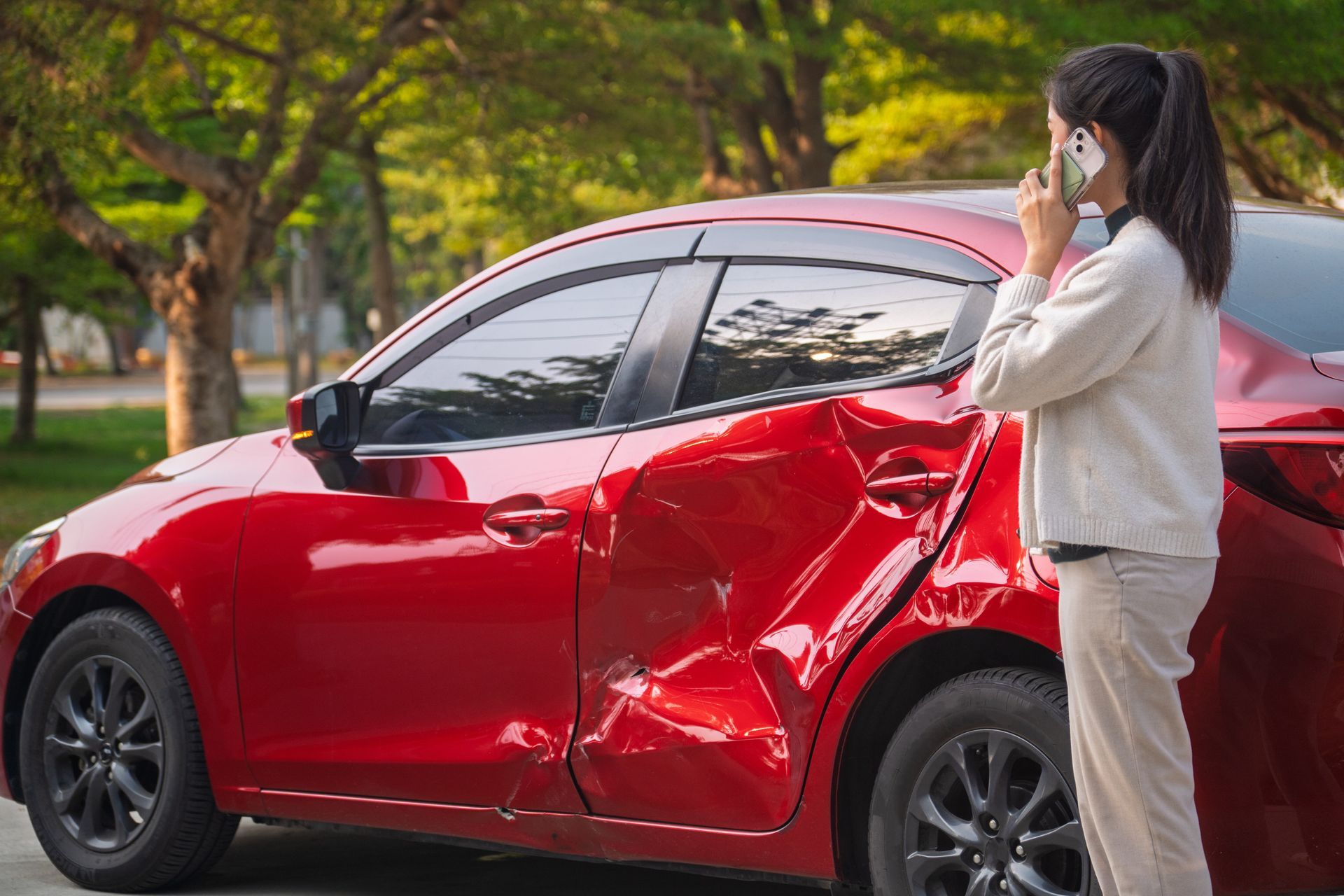 Woman on phone beside a red car with significant damage on the side, likely after a collision.