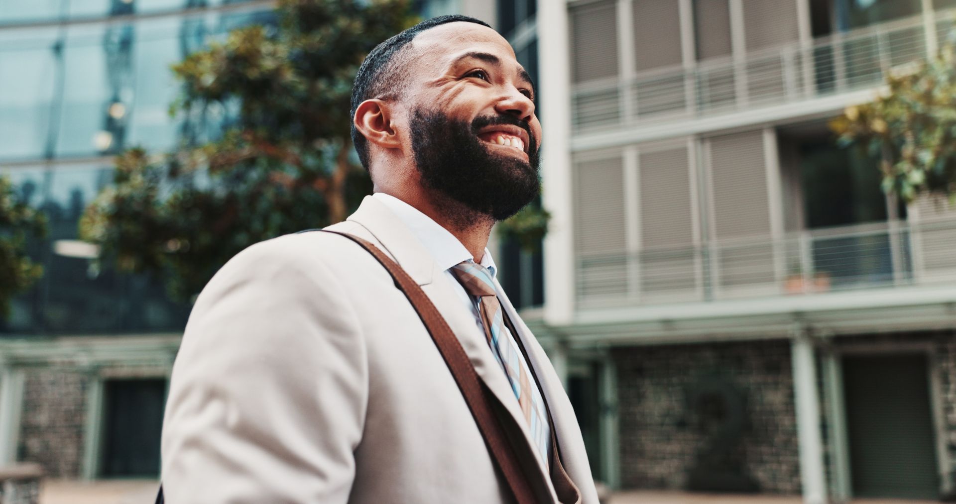 Man in suit smiles, carrying bag, walking outside near modern building.