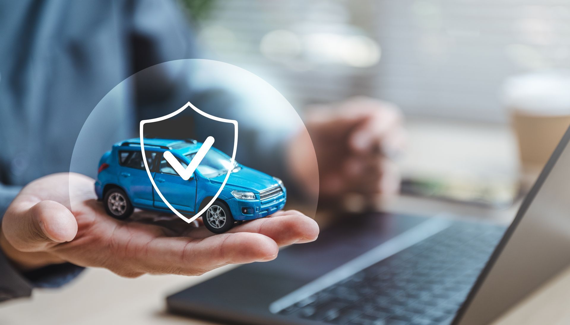 Hand holding a blue toy car with a shield and checkmark overlay near a laptop.