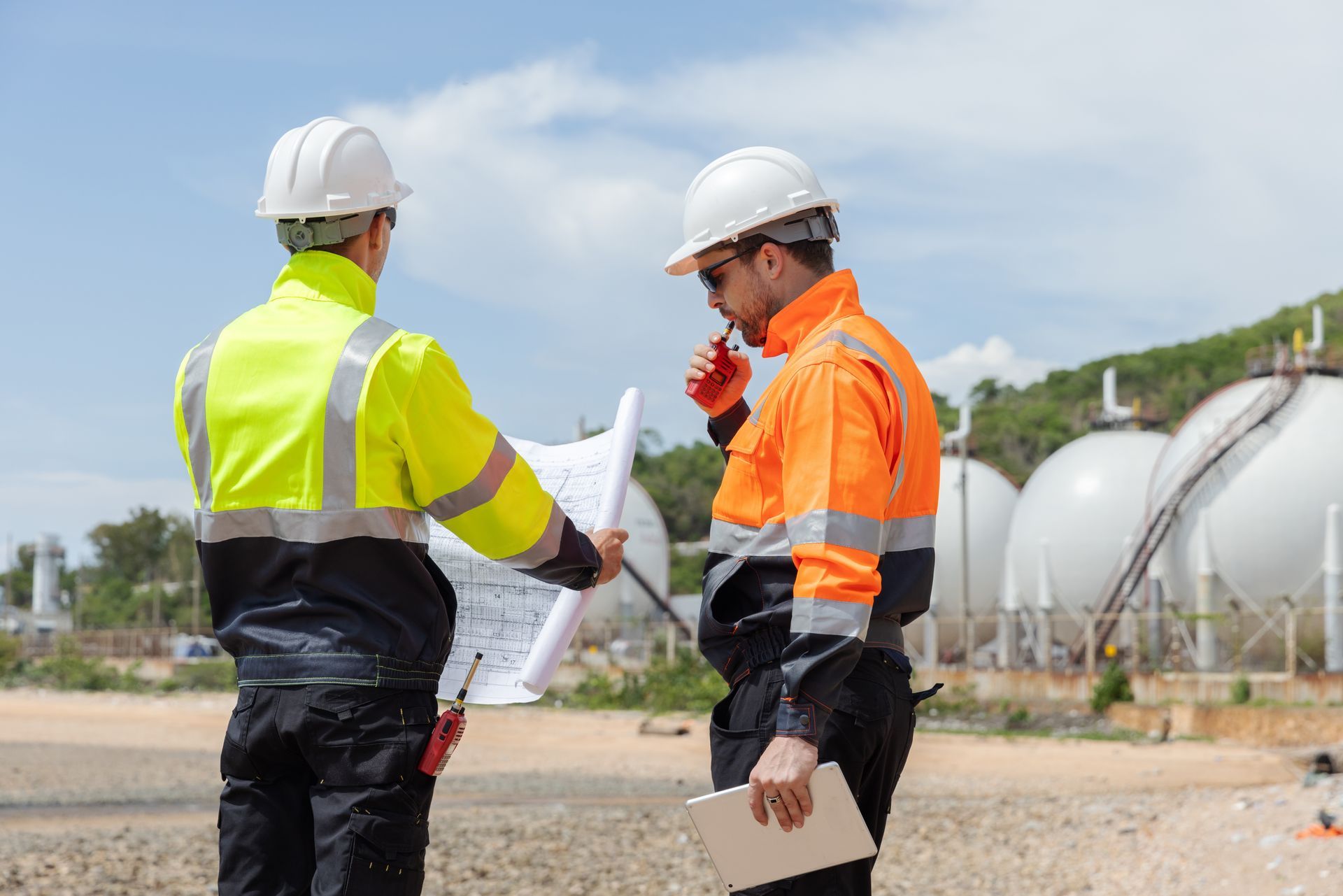 Two construction workers reviewing blueprints near industrial tanks. One holds a radio, the other a tablet.
