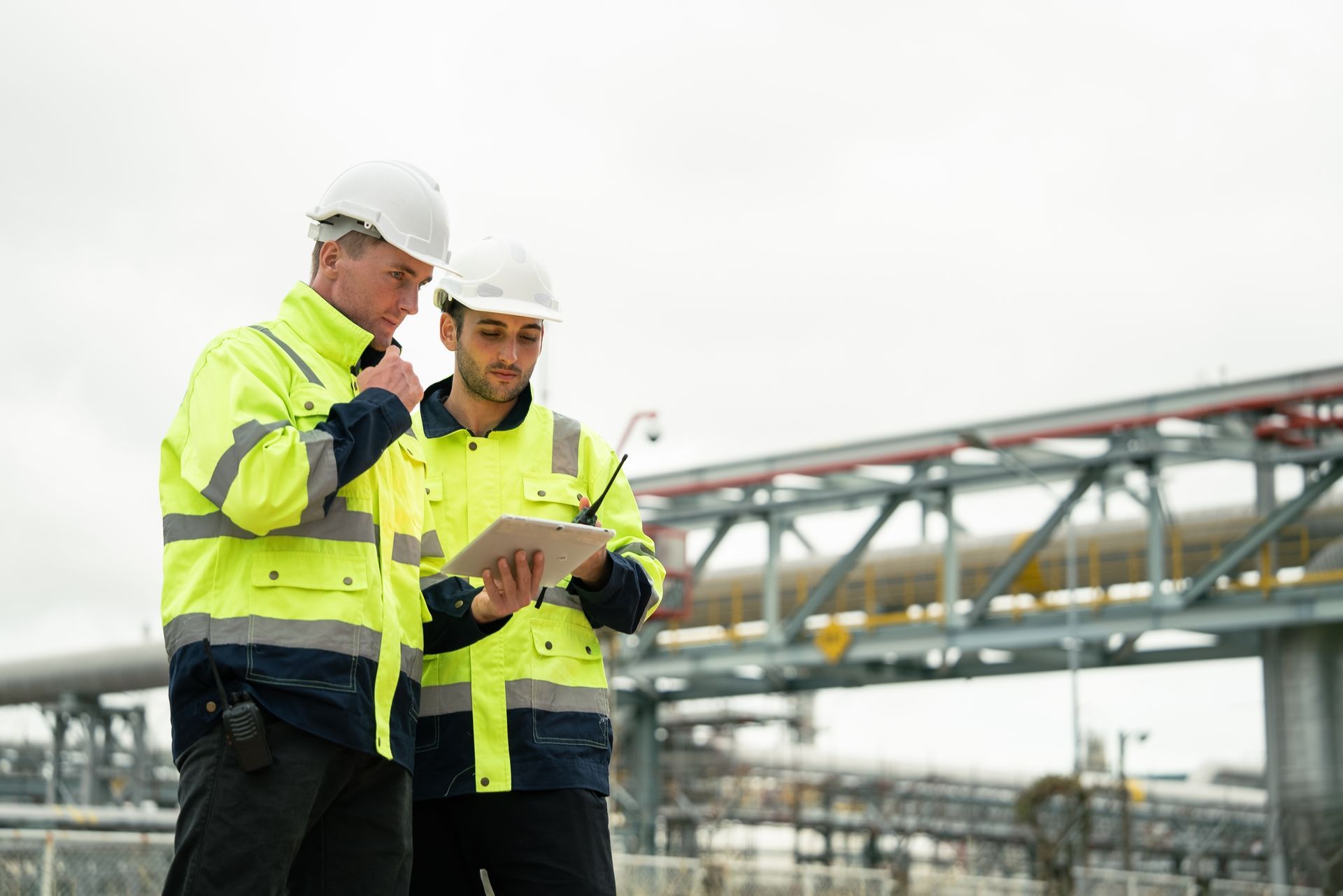 Two engineers in hard hats and safety vests review a tablet outdoors near industrial equipment.