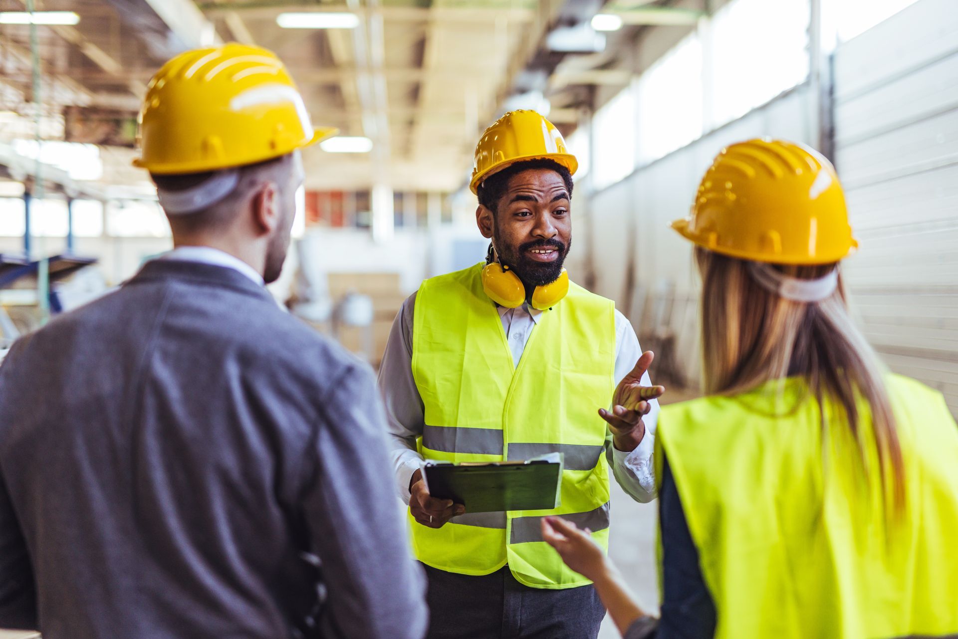 Three people in a warehouse, wearing hard hats and safety vests, discussing.