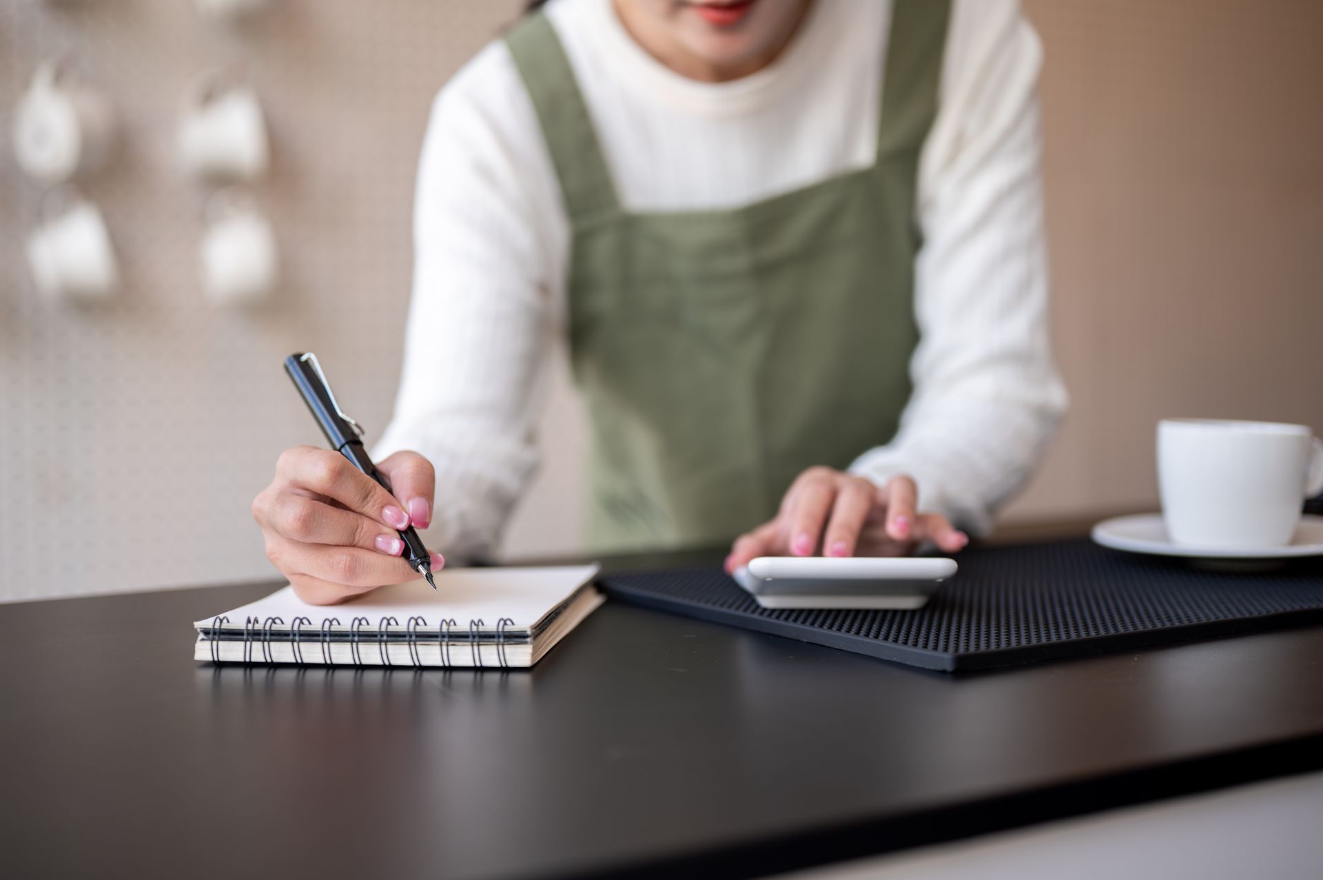 Person writing in a notebook, with a calculator and coffee cup on a black counter.