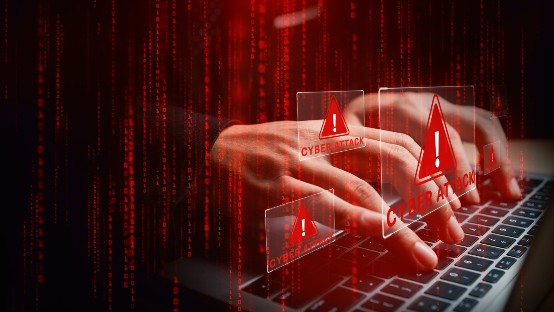 Person's hands typing on a laptop with red alert graphics overlaying the screen, set against a dark background with digital code.