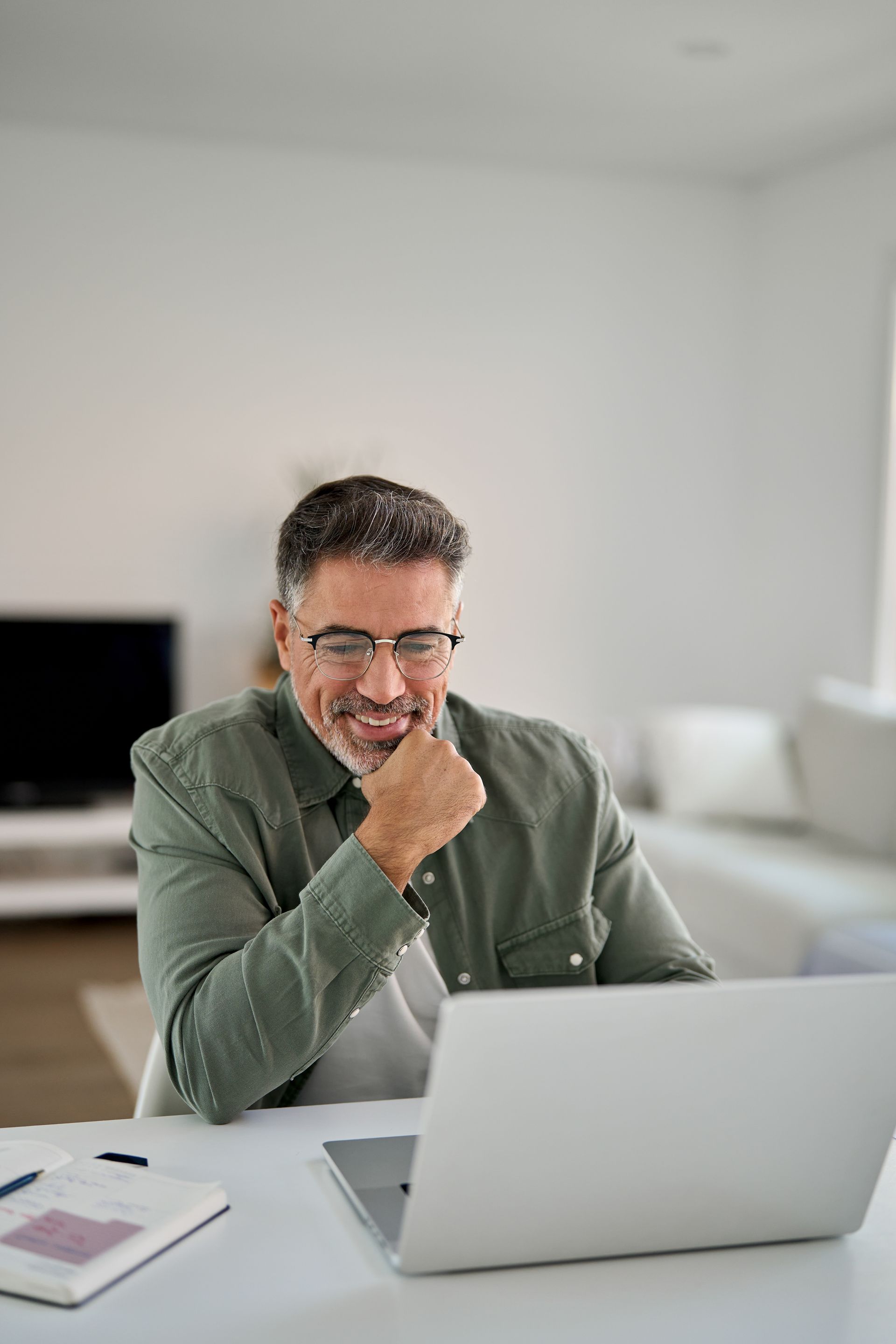 Man smiles while looking at a laptop, seated at a table. He has short graying hair and is wearing glasses and a green shirt.