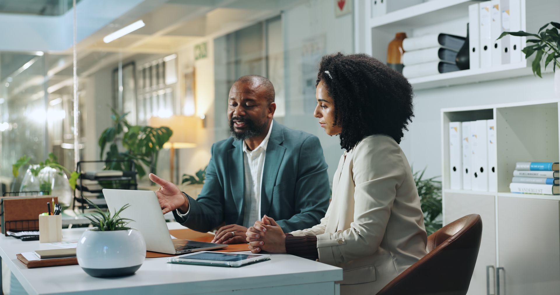 Man points to laptop screen, discussing with woman at desk in modern office.