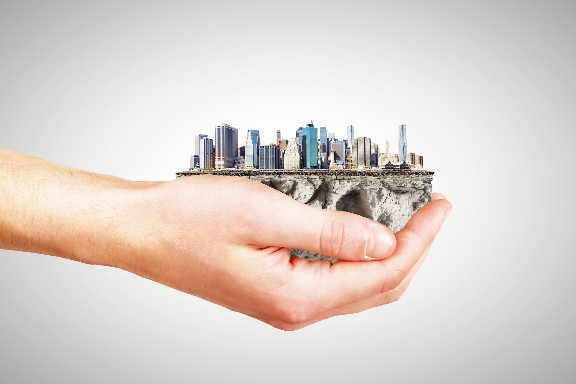 Hand holding a miniature city on a piece of land, with tall buildings and a light gray background.