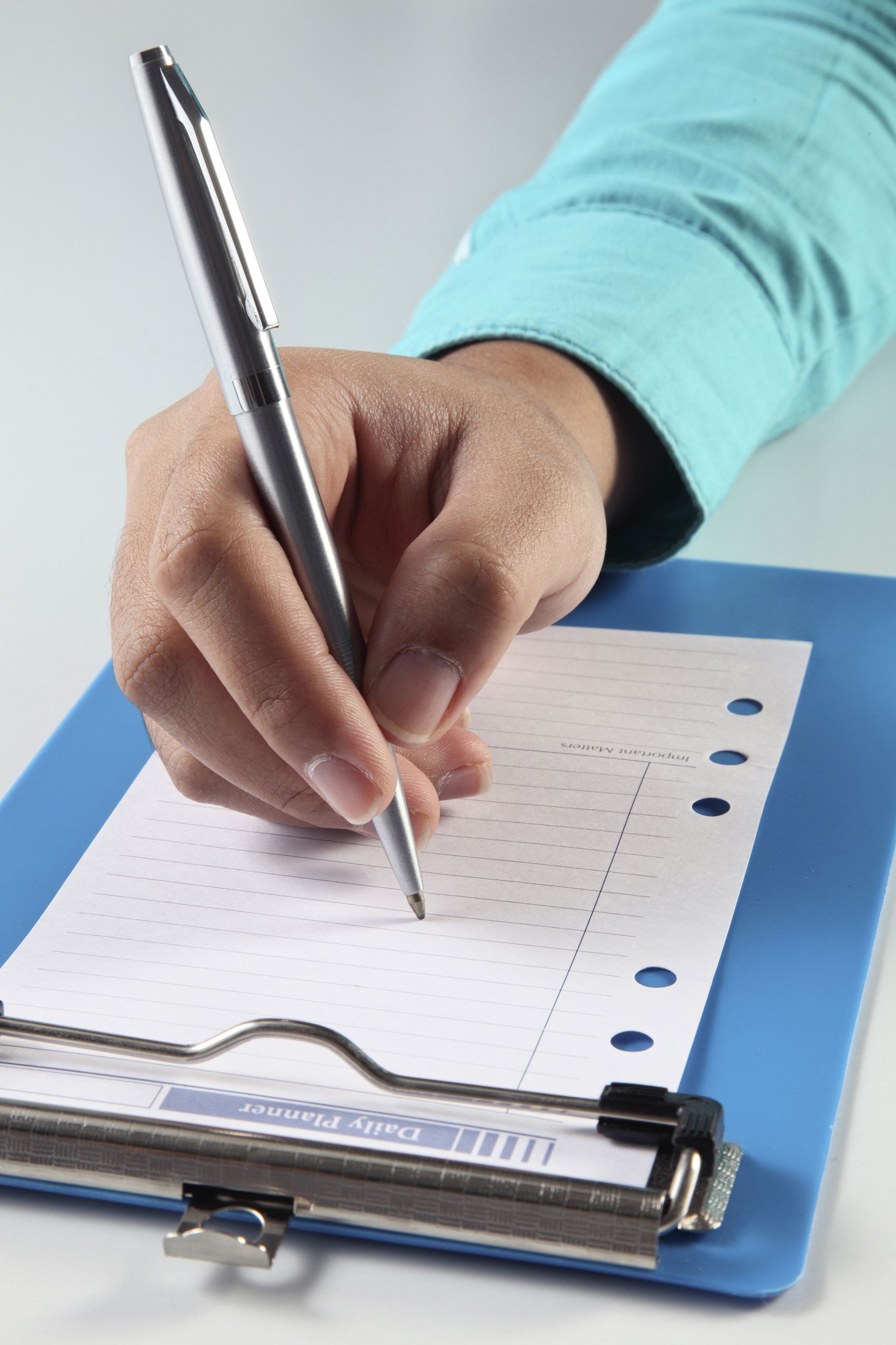 Person's hand holding a pen, writing on a dotted paper sheet held by a blue clipboard. Turquoise sleeve visible.