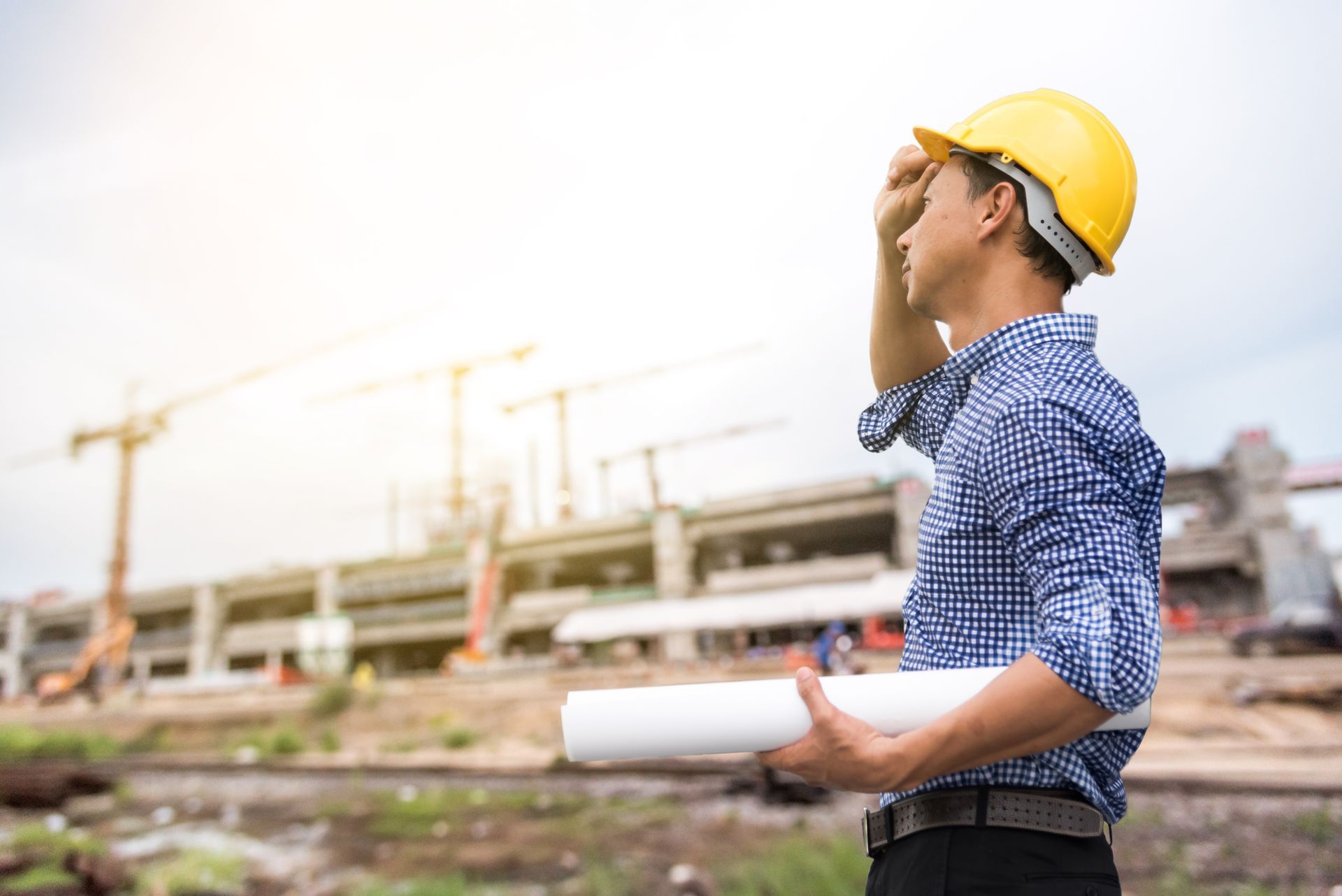 Construction worker in yellow hard hat holding plans, looking at a construction site on a sunny day.