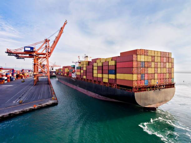 Cargo ship docked at a port, loaded with containers, near an orange crane and blue water.