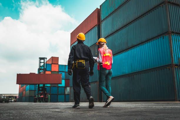 Two people in safety vests and hard hats at a shipping port, surrounded by cargo containers.
