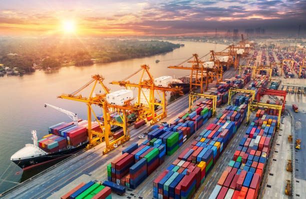 Cargo ship at a port being loaded/unloaded by cranes; shipping containers in rows, sunset.