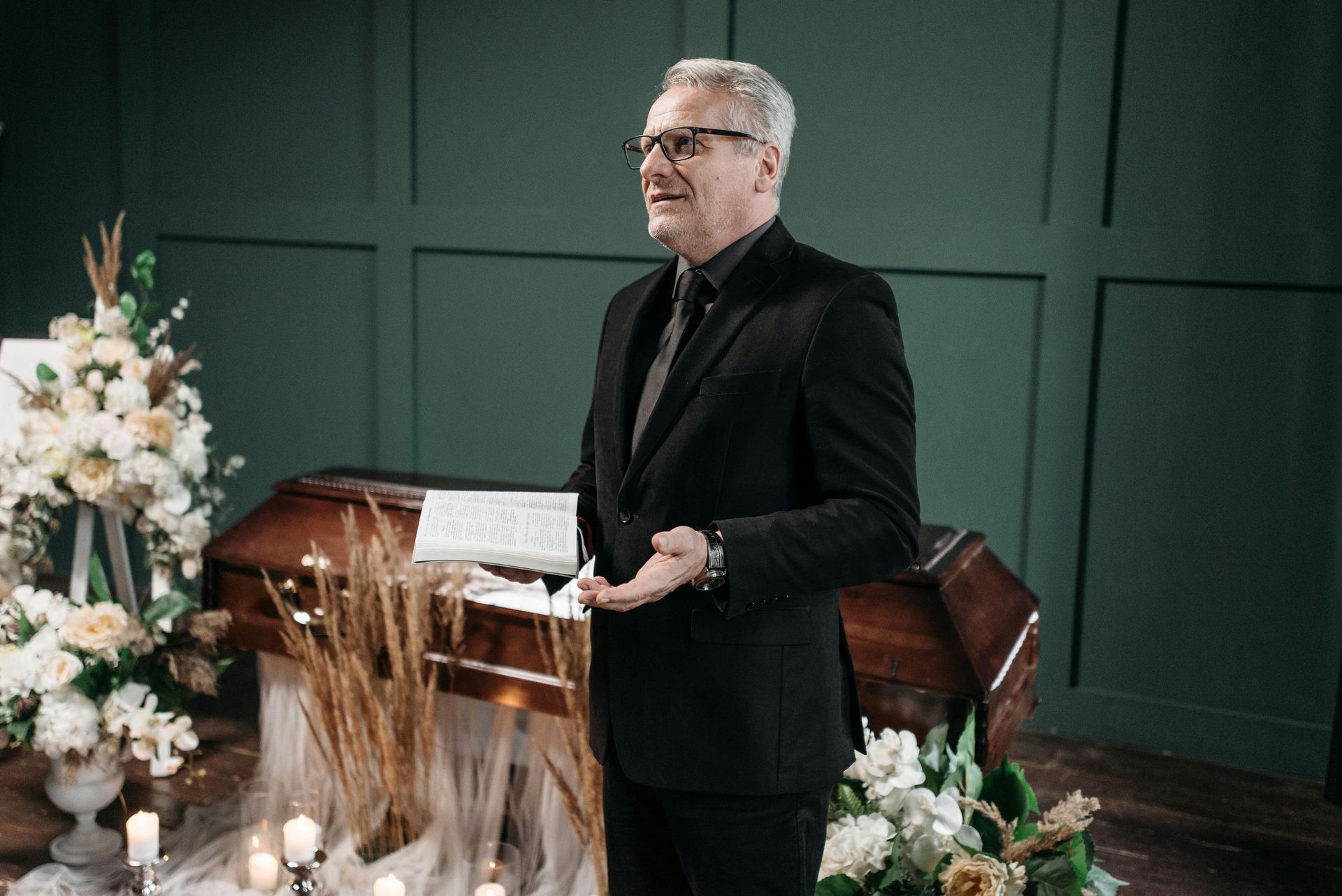 Man in a black suit speaking at a podium beside a wooden casket and floral arrangements