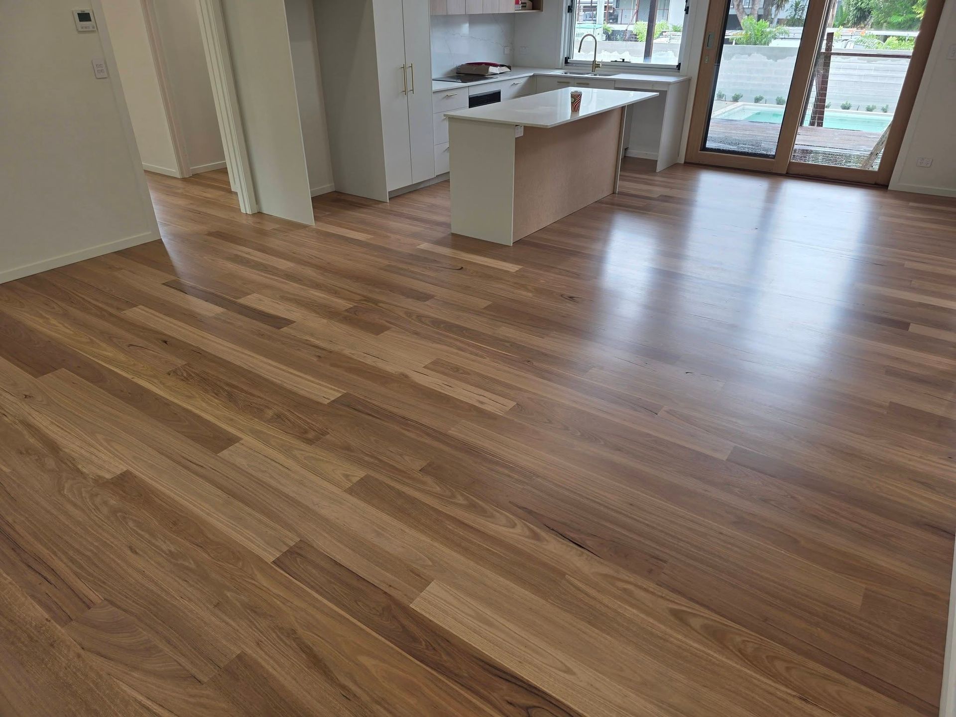Wooden flooring in a new house, near a kitchen island, and glass doors. Shiny and brown — Alan Aldridge Floor Sanding in Urraween, QLD