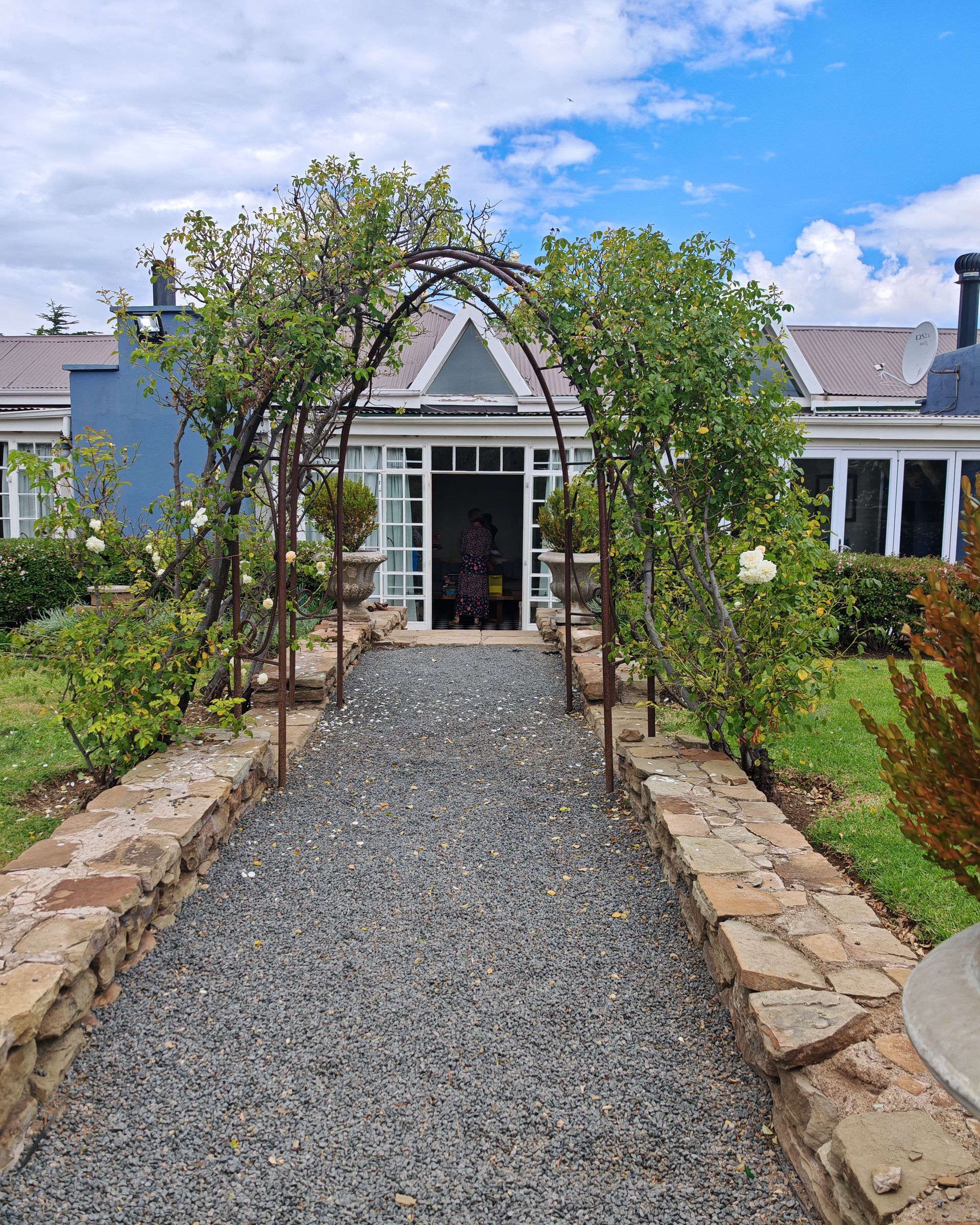 Gravel walkway leading to a white house entrance framed by arching trees and stone borders