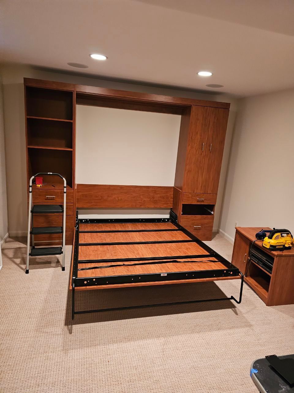 A brown, wooden Murphy bed frame and storage cabinets in a beige-carpeted room.