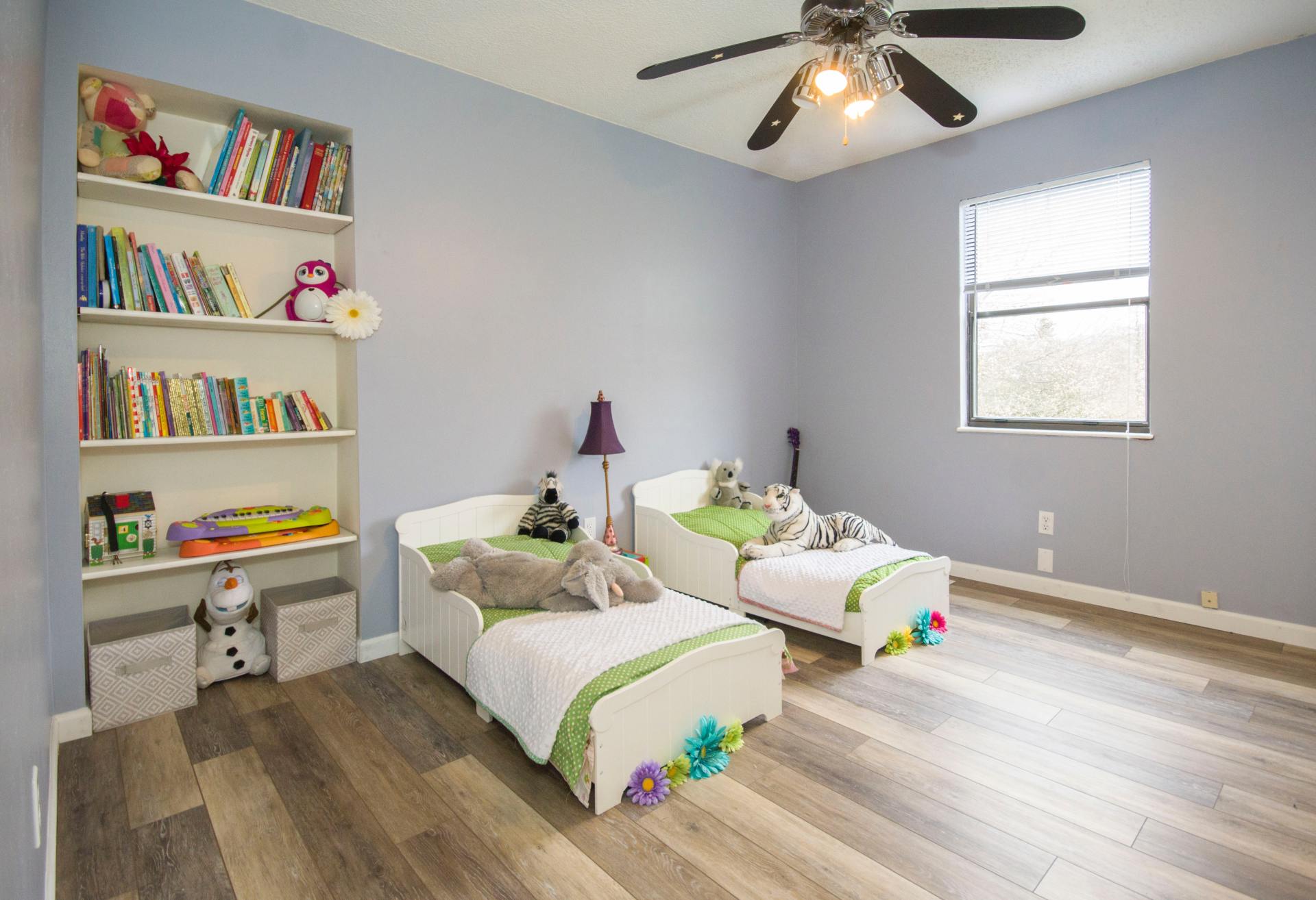 Child's bedroom with two beds, built-in bookshelf, and a window. Light blue walls, wood-look floor.