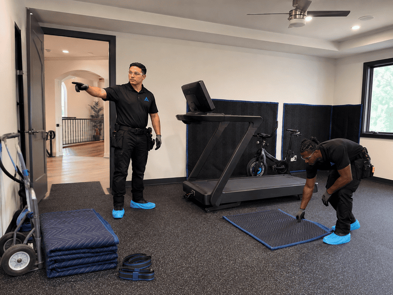 Two workers setting up a treadmill in a bright home gym, with mats and equipment nearby.