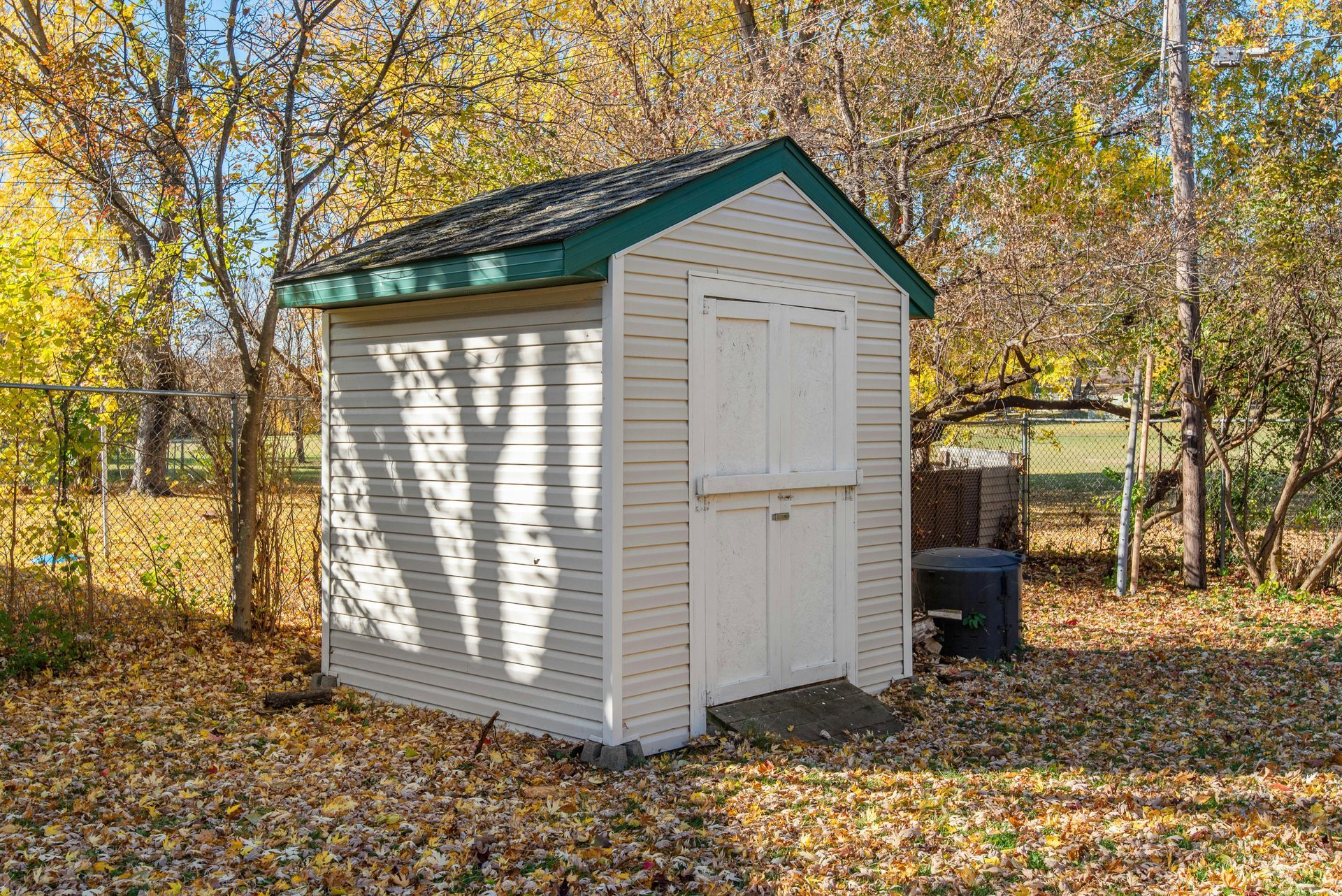 Small white shed with green trim in a yard with fall leaves.