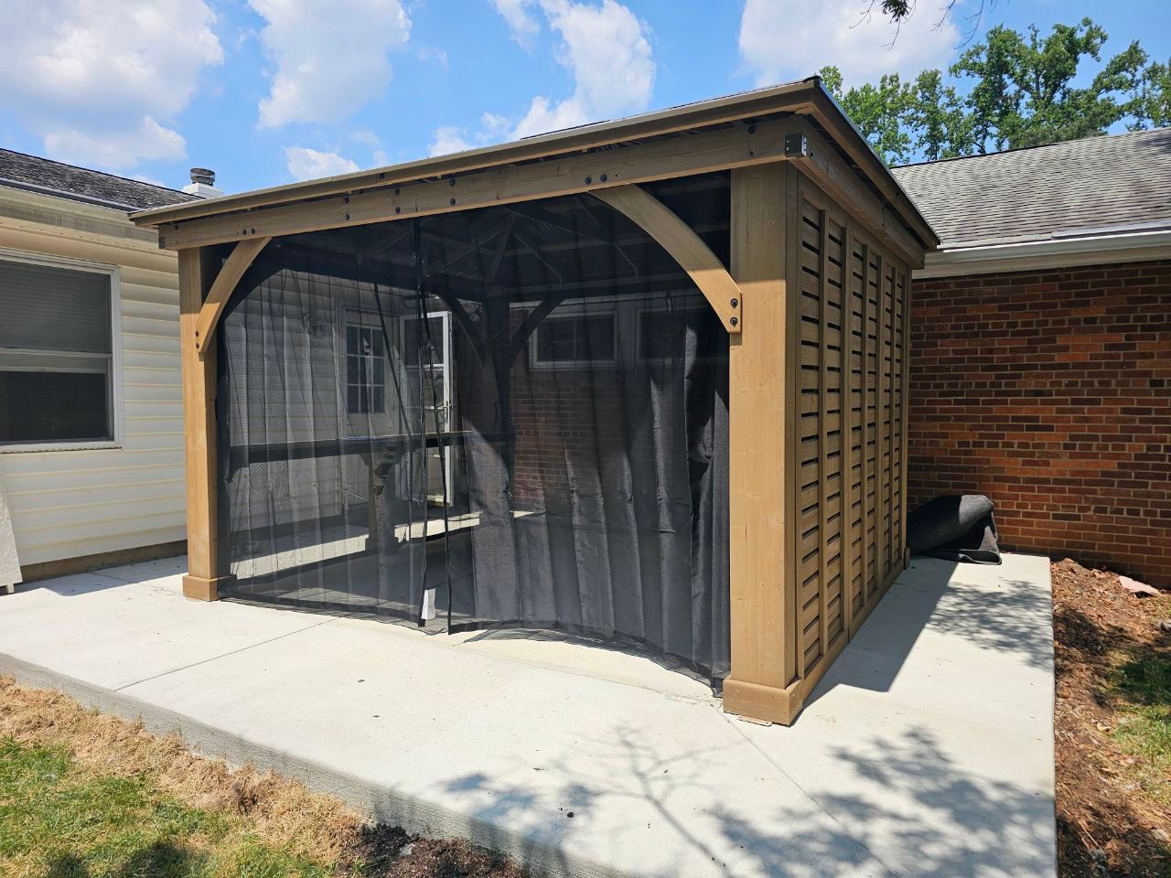Brown gazebo with mesh curtains on a concrete patio next to a brick building.