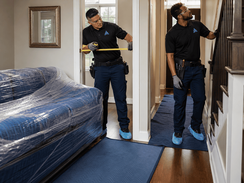 Two movers carrying furniture through a hallway, one holding a wrapped sofa and one near a doorway.