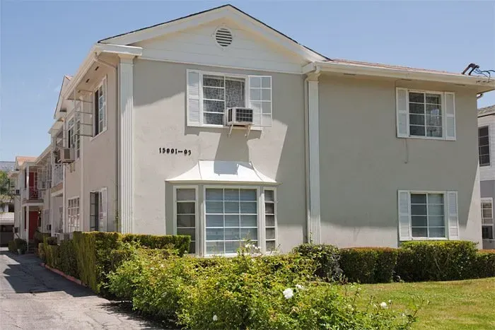 Two-story beige apartment building with white trim, green hedges, and a small parking area beside it