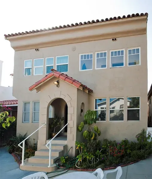 Two-story beige house with red-tiled entrance canopy and white-framed windows, front steps, and garden shrubs