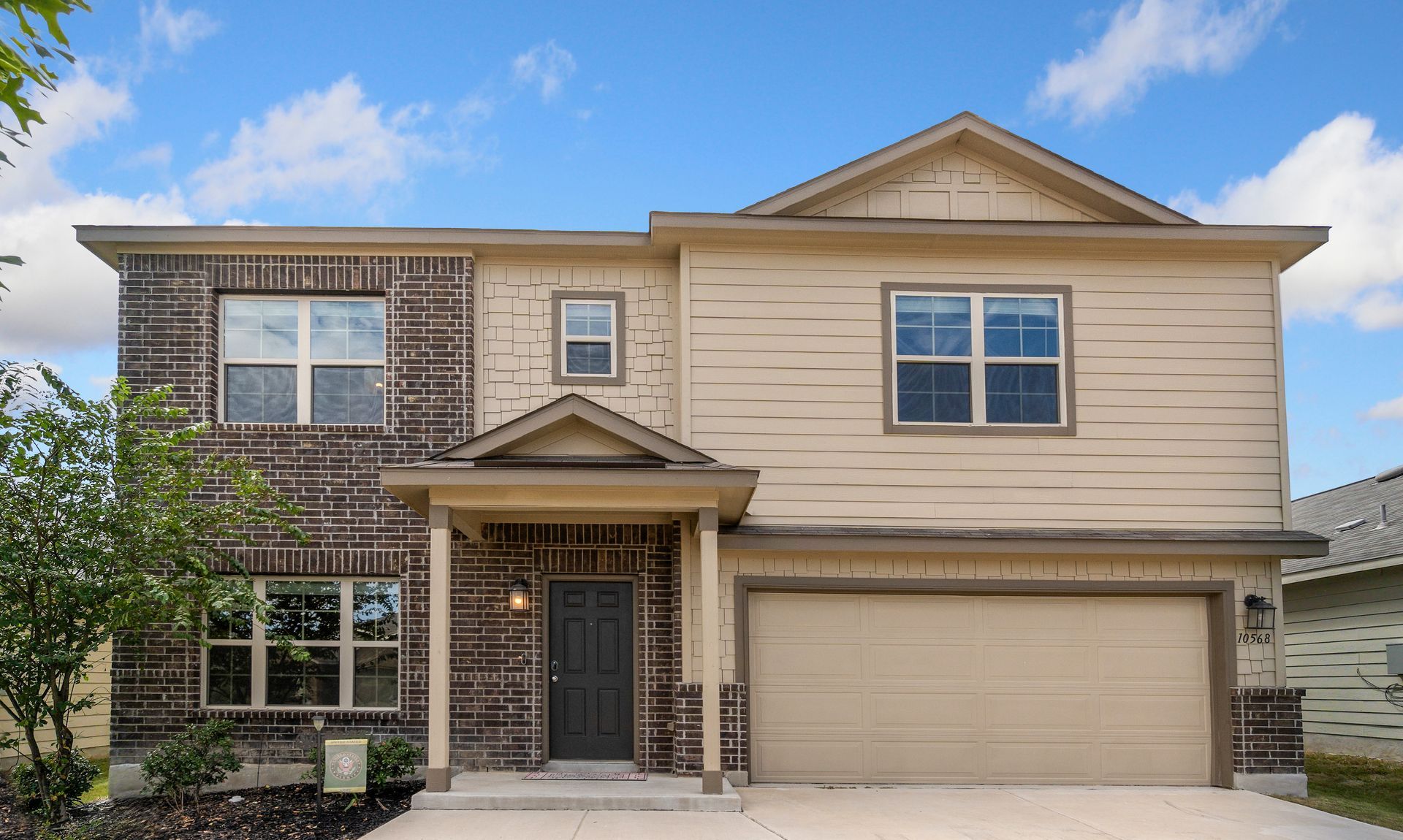 Two-story beige suburban house with a brick facade and two-car garage under a blue sky