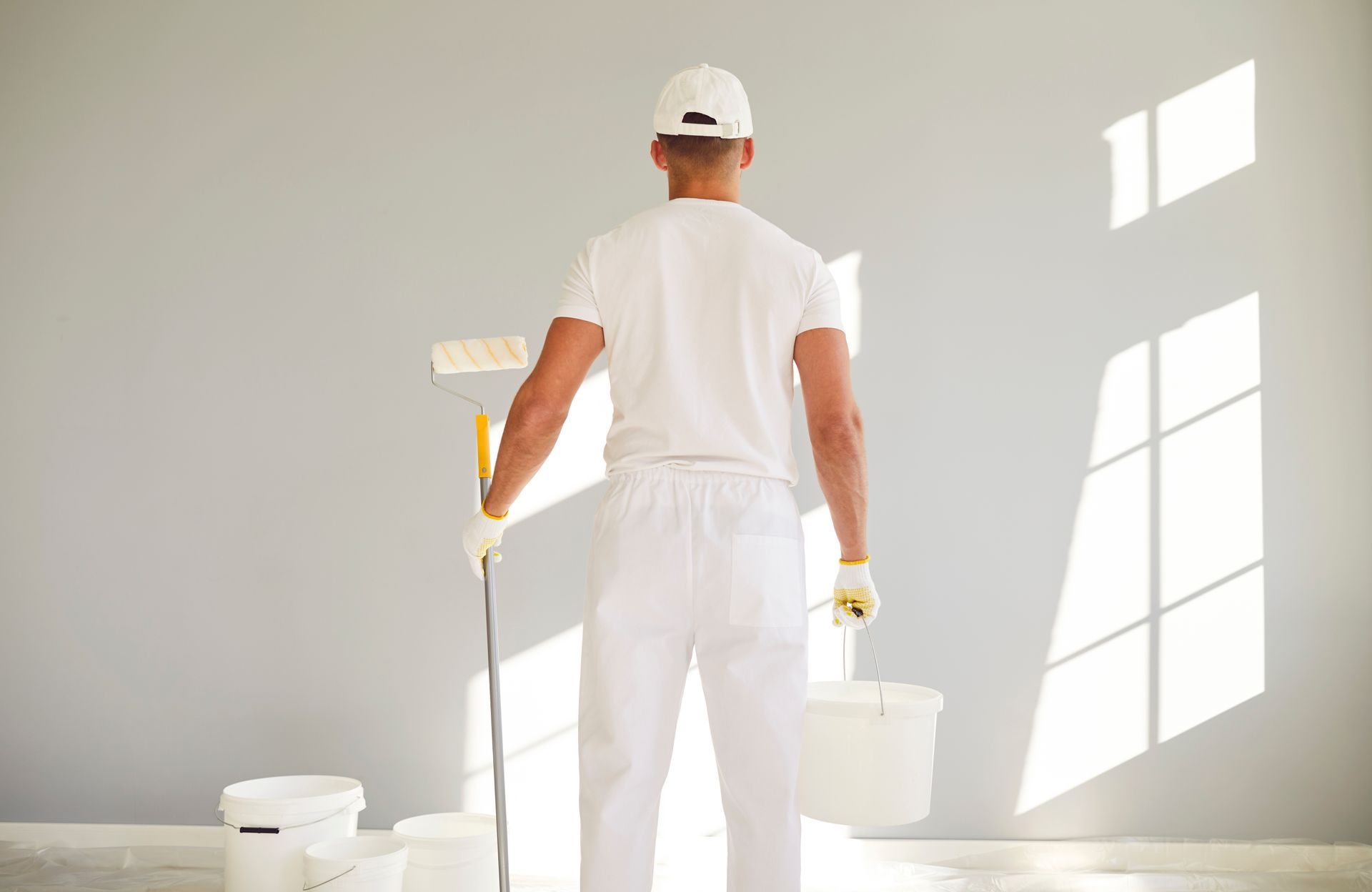 Painter in white workwear holding a bucket and brush in a bright room with fresh white walls. Painter in white workwear holding a bucket and brush in a bright room with fresh white walls.