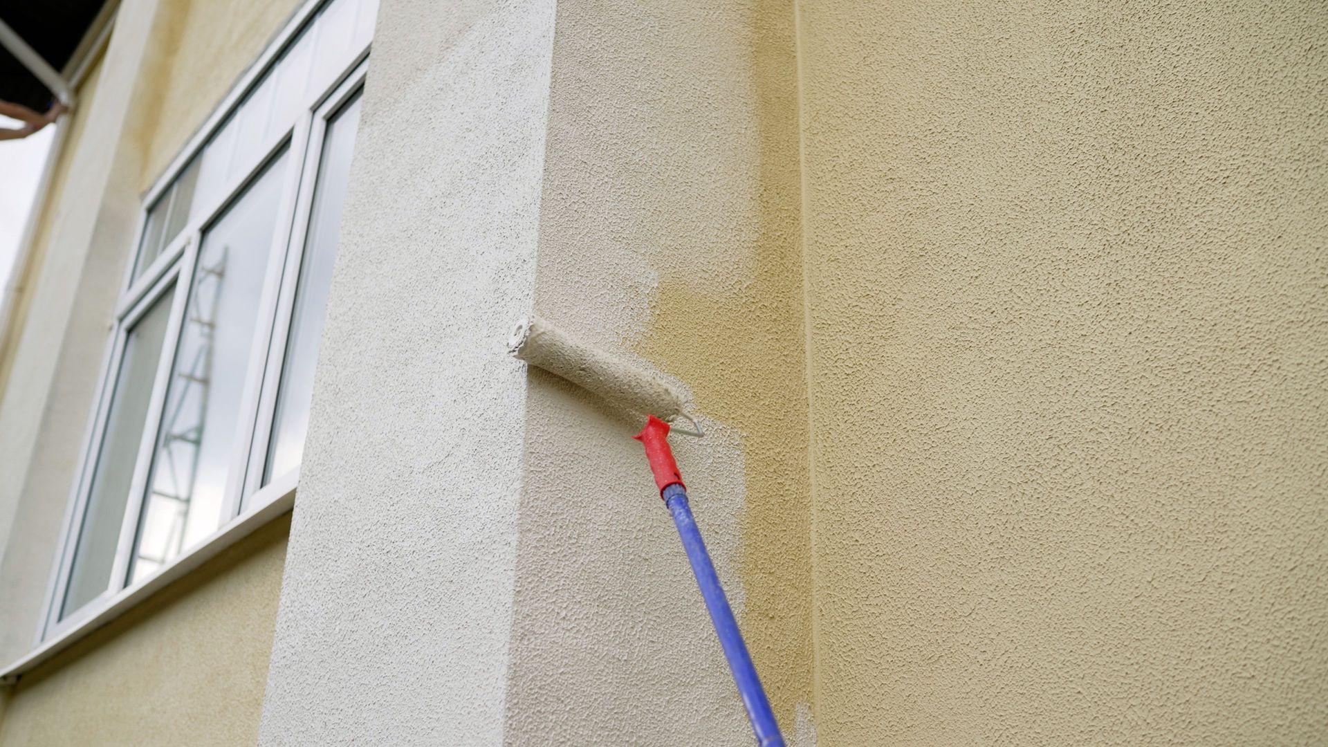 Paint roller applying red paint to a building corner wall next to a window