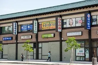Street view of a Korean storefront with signs, shuttered ground-floor shops, and two pedestrians walking by